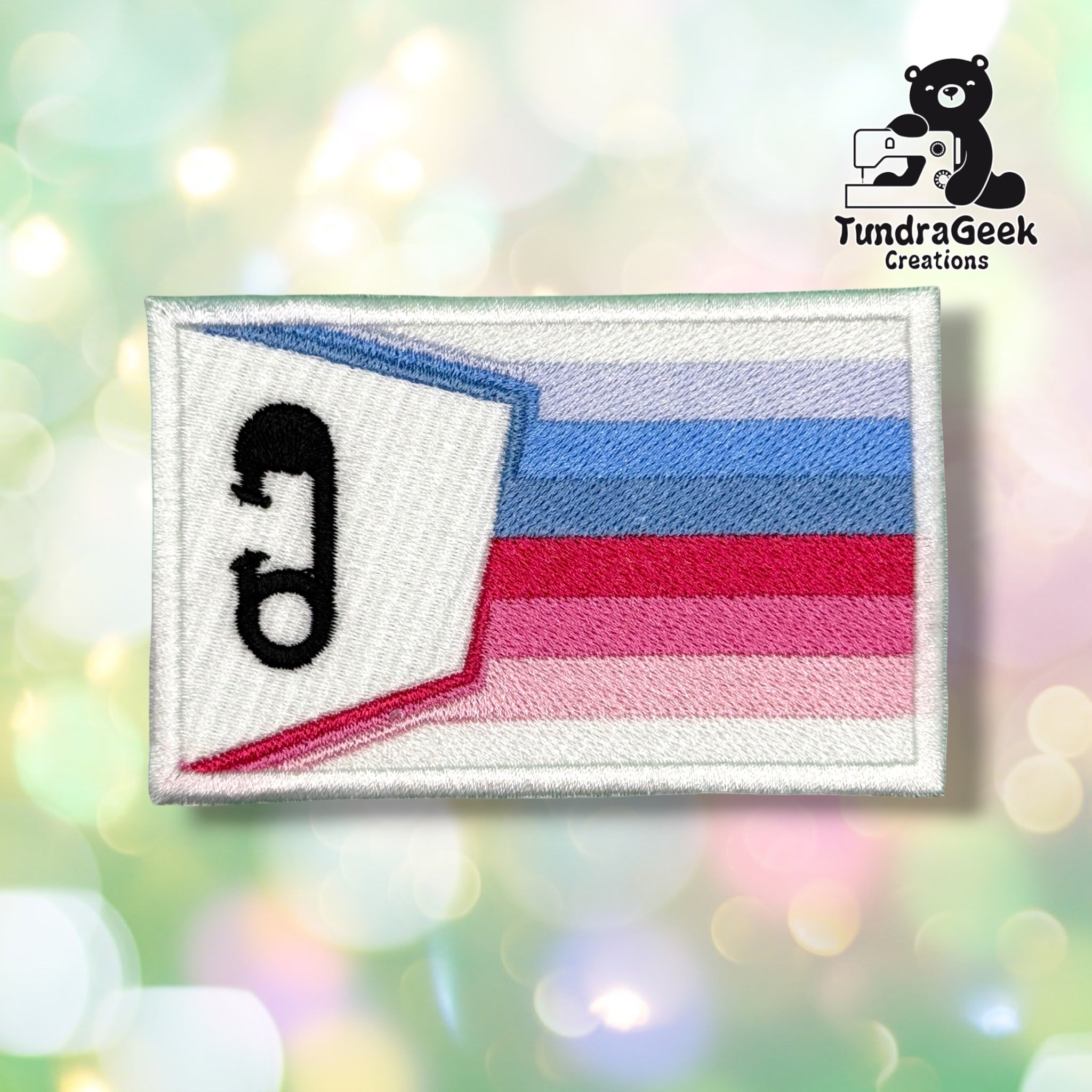 Front view of a rectangular white satin patch embroidered with a small waving ABDL pride flag graphic — light blue, blue, white, magenta, pink, and light pink horizontal stripes — with a black embroidered diaper pin icon on the flag's white fold.