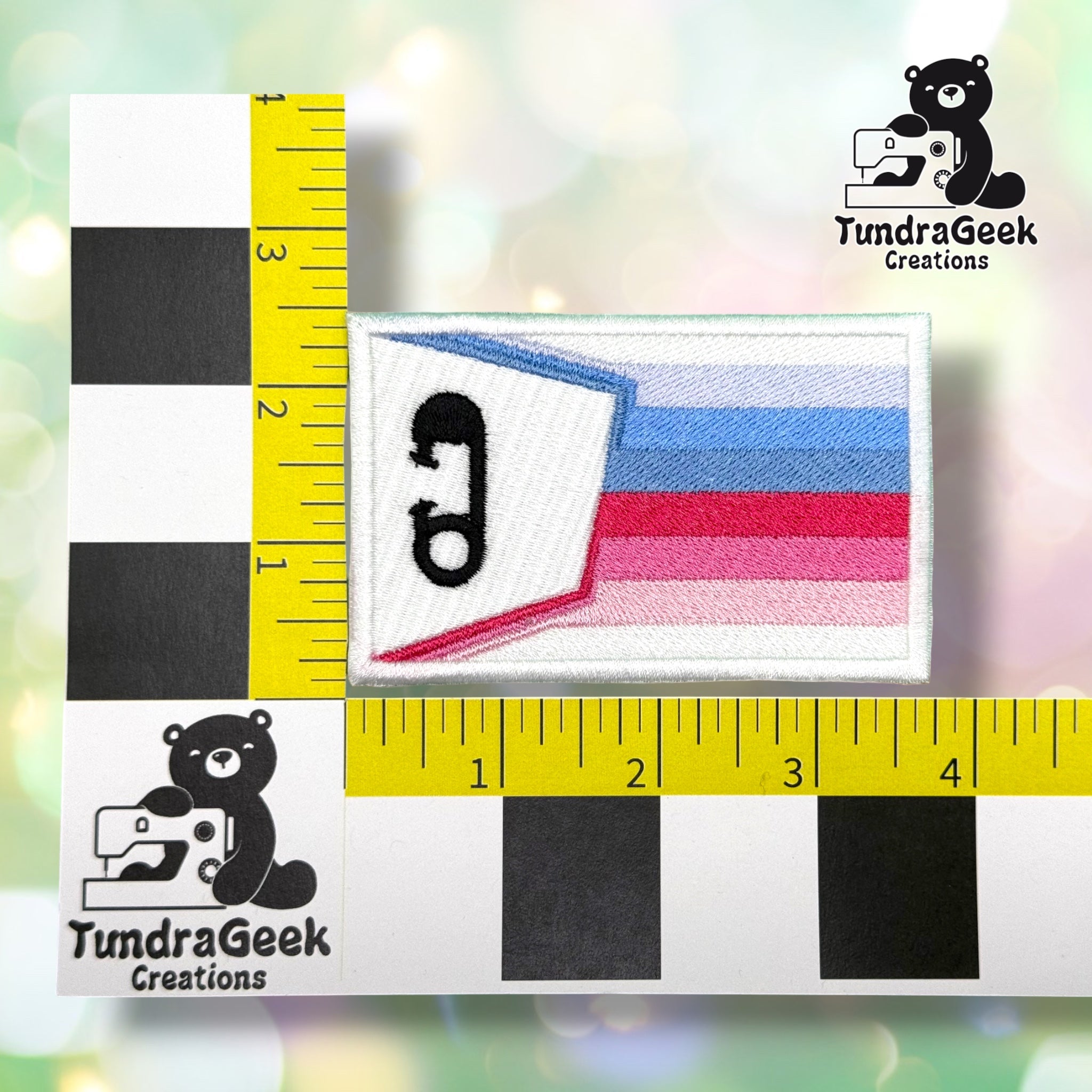 Patch shown against a black-and-white checkered background with yellow rulers for scale, measuring approximately 3.75 inches wide by 2.25 inches tall.  ---