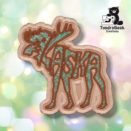 Front view of a patch shaped around the word "ALASKA," formed into the silhouette of a standing moose with antlers, legs, and a tail. Embroidered in tan, brown, and green thread on a tan twill background.