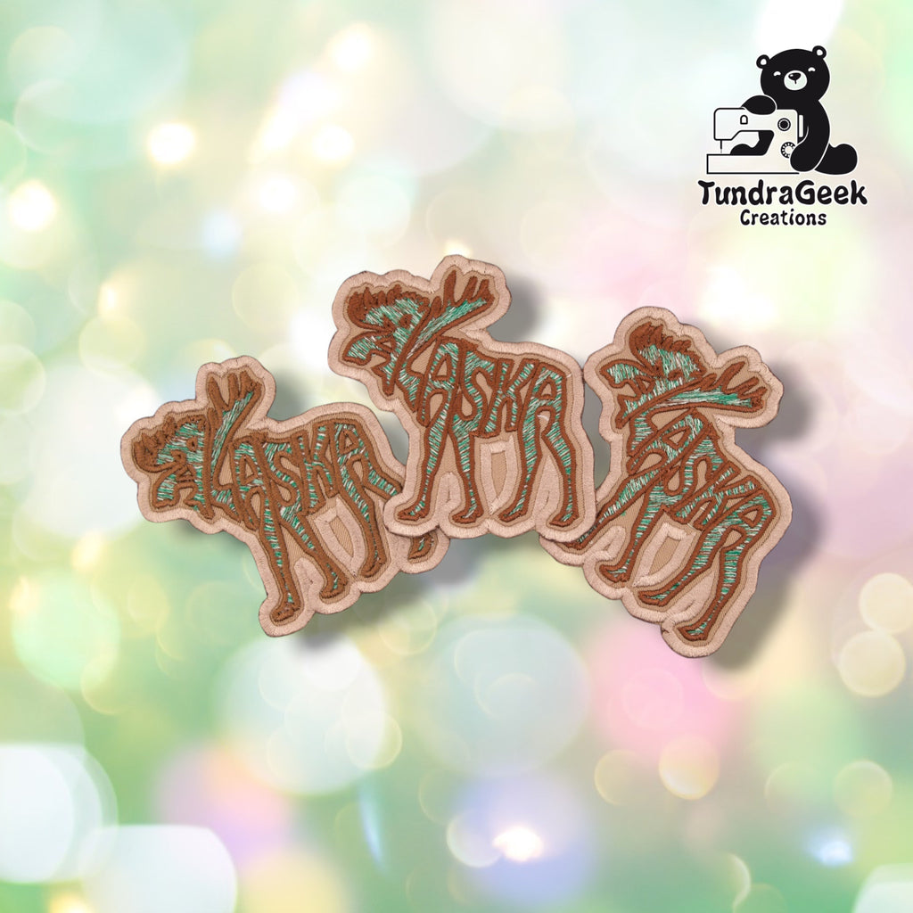 Three identical Alaska Moose patches fanned out together, each showing the same word-art moose design in tan, brown, and green.