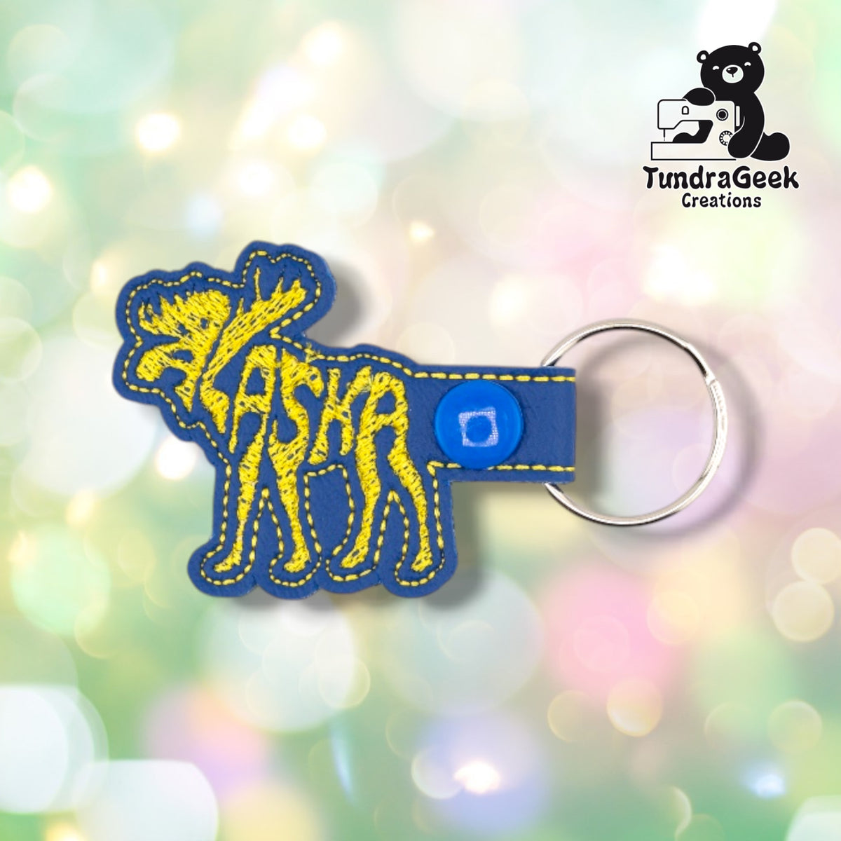 Front of a blue vinyl keychain, with the word "ALASKA" embroidered in yellow thread, shaped into the silhouette of a standing moose with antlers and legs. Blue plastic snap closure and metal split ring at top.