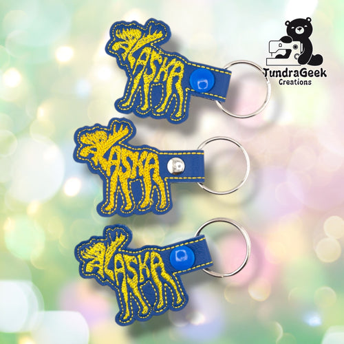 Three identical Alaskan Moose keychains fanned out together, each showing the same blue vinyl body and yellow word-art moose design.