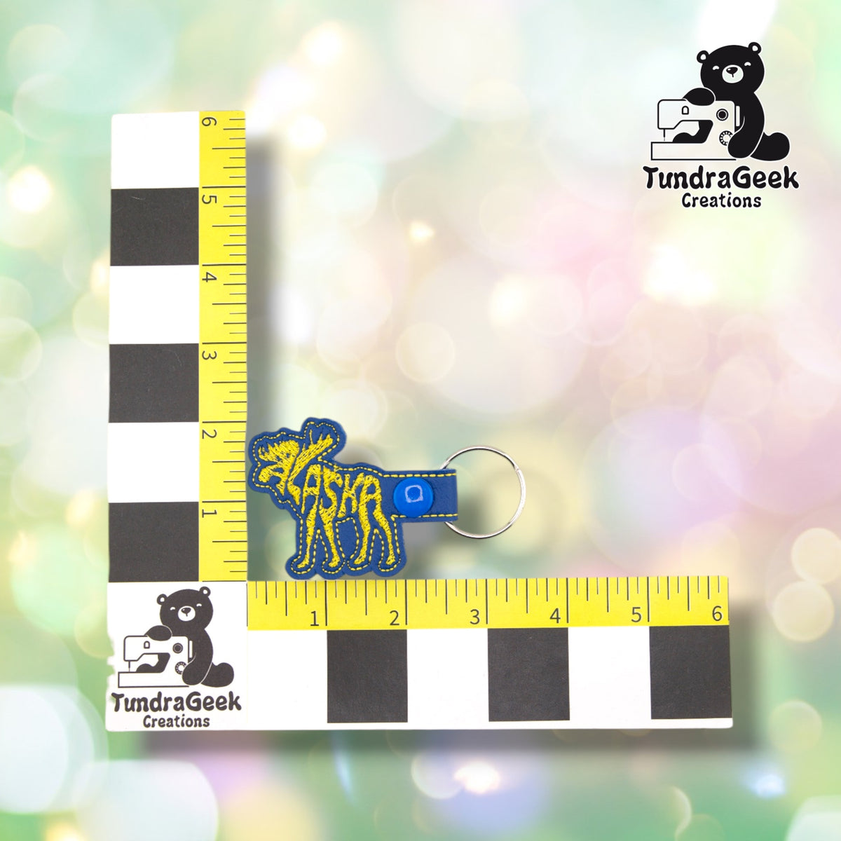 Alaskan Moose keychain shown against a black-and-white checkered background with a yellow ruler for scale, measuring approximately 3.5 inches wide by 2.25 inches tall.  ---