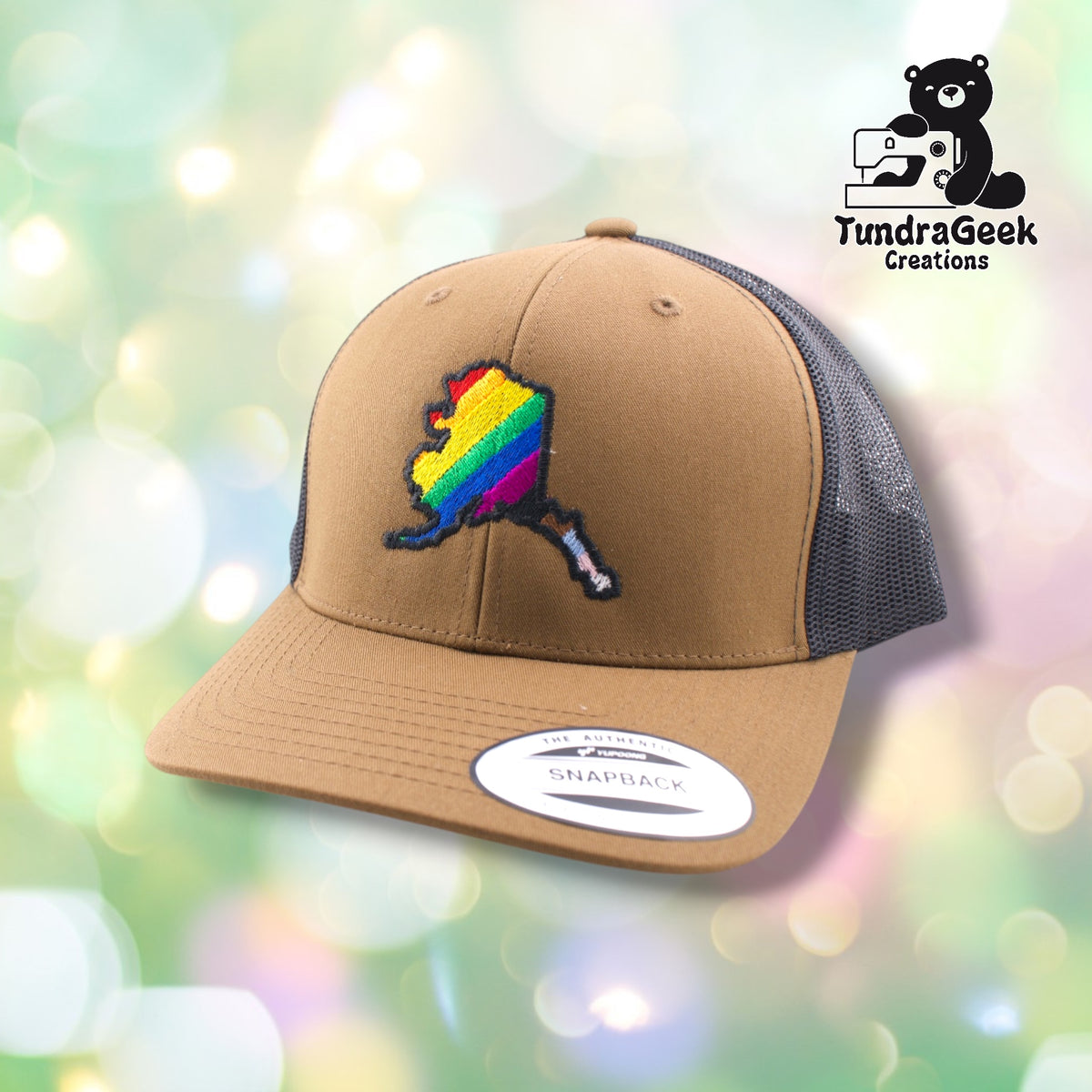 A tan and gray snapback trucker cap with a rainbow-colored embroidered Alaska state map design on the front panel.
