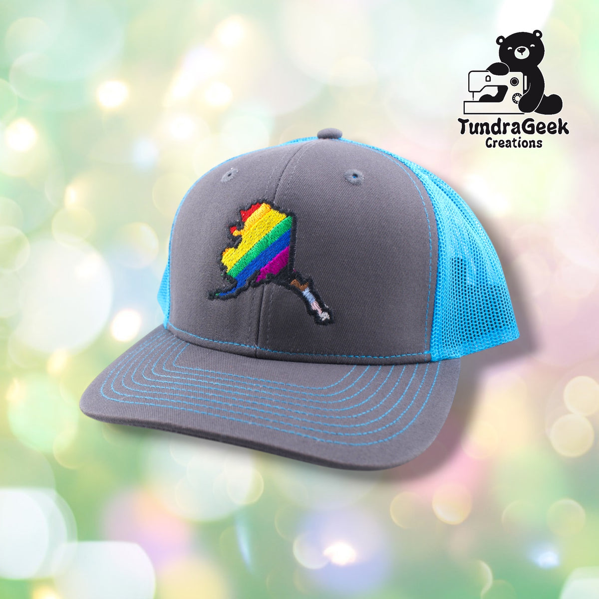 A gray and turquoise trucker cap with rainbow-striped Alaska state embroidery on the front, displayed against a soft bokeh background.