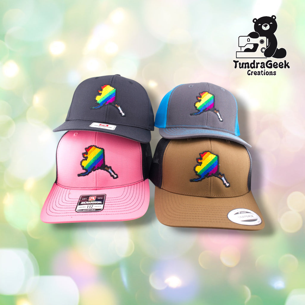 Four baseball caps in black, pink, gray-blue, and tan featuring embroidered rainbow Alaska state designs displayed against a soft bokeh background.