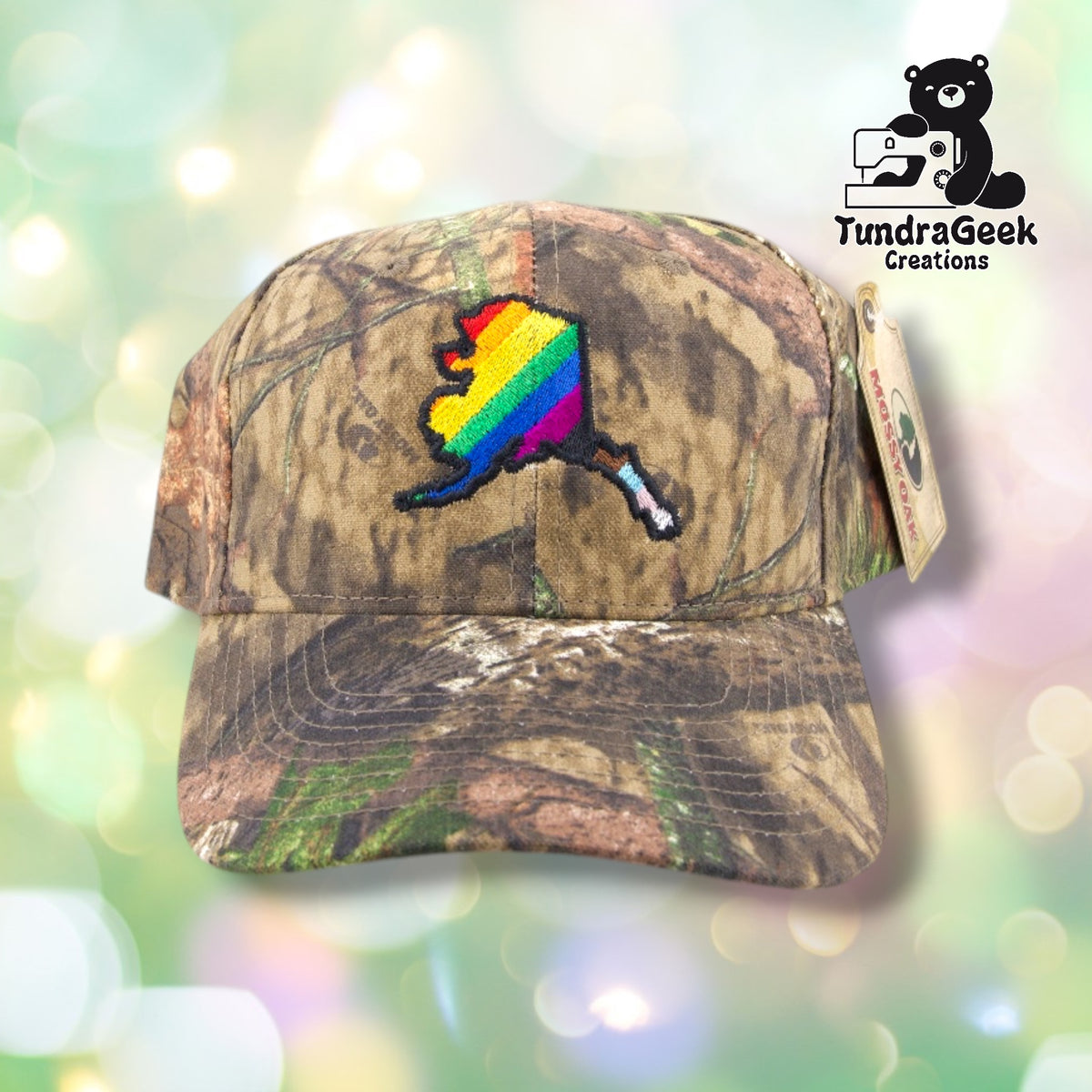 A camouflage baseball cap with a rainbow-colored embroidered Alaska state shape on the front, displayed against a soft-focus background.