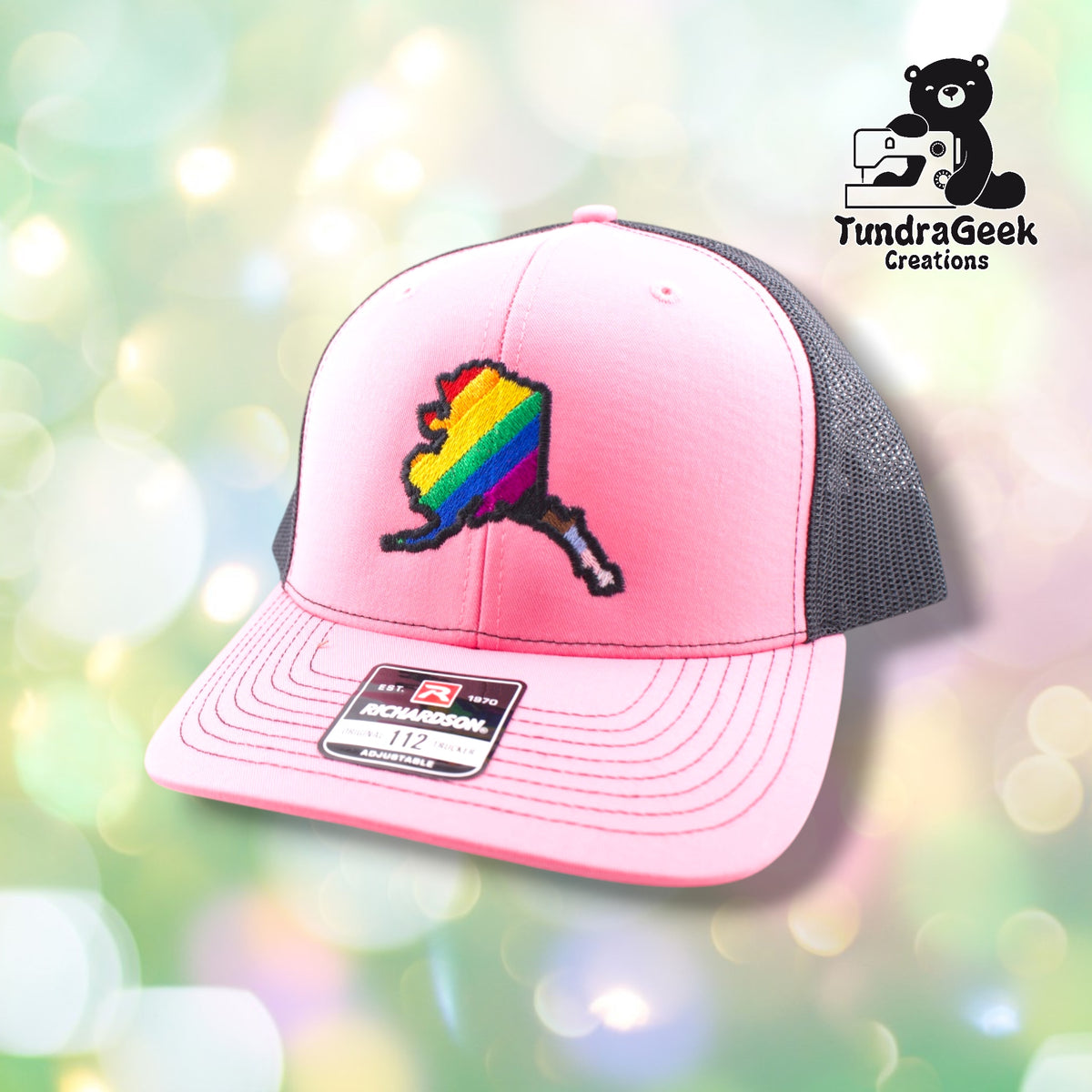 A pink and gray trucker cap with a rainbow-striped Alaska state emblem on the front and an adjustable back patch visible.