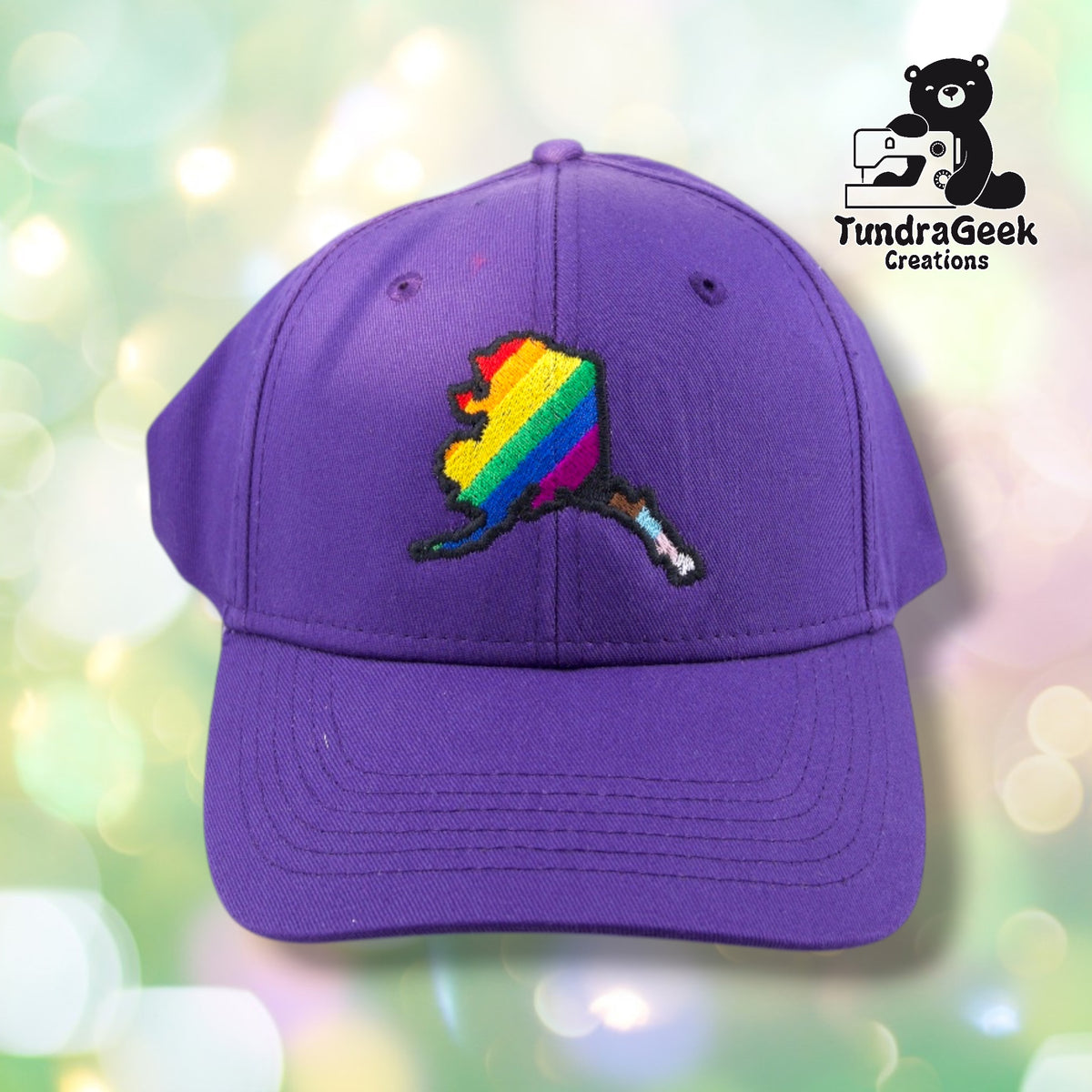 A purple baseball cap with an embroidered rainbow map of Alaska and brown handle, displayed against a soft bokeh background.