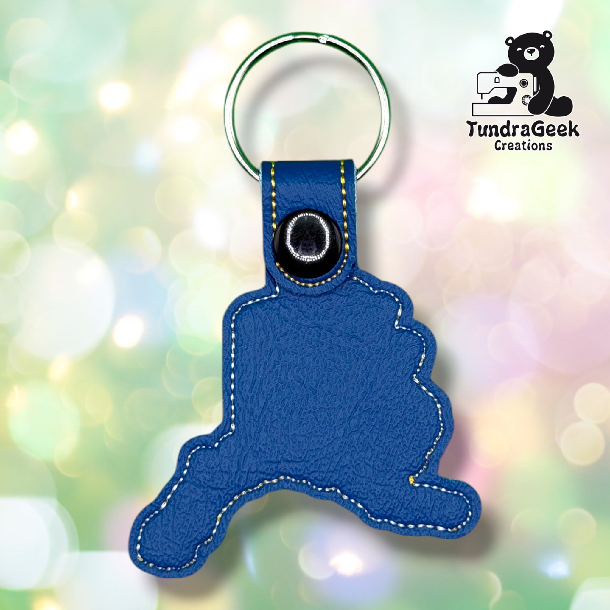 Back of a blue vinyl keychain shaped like the state of Alaska, with white and gold topstitched outline, a black plastic snap closure at the top of the tab, and a silver split ring. No embroidery visible from this side.
