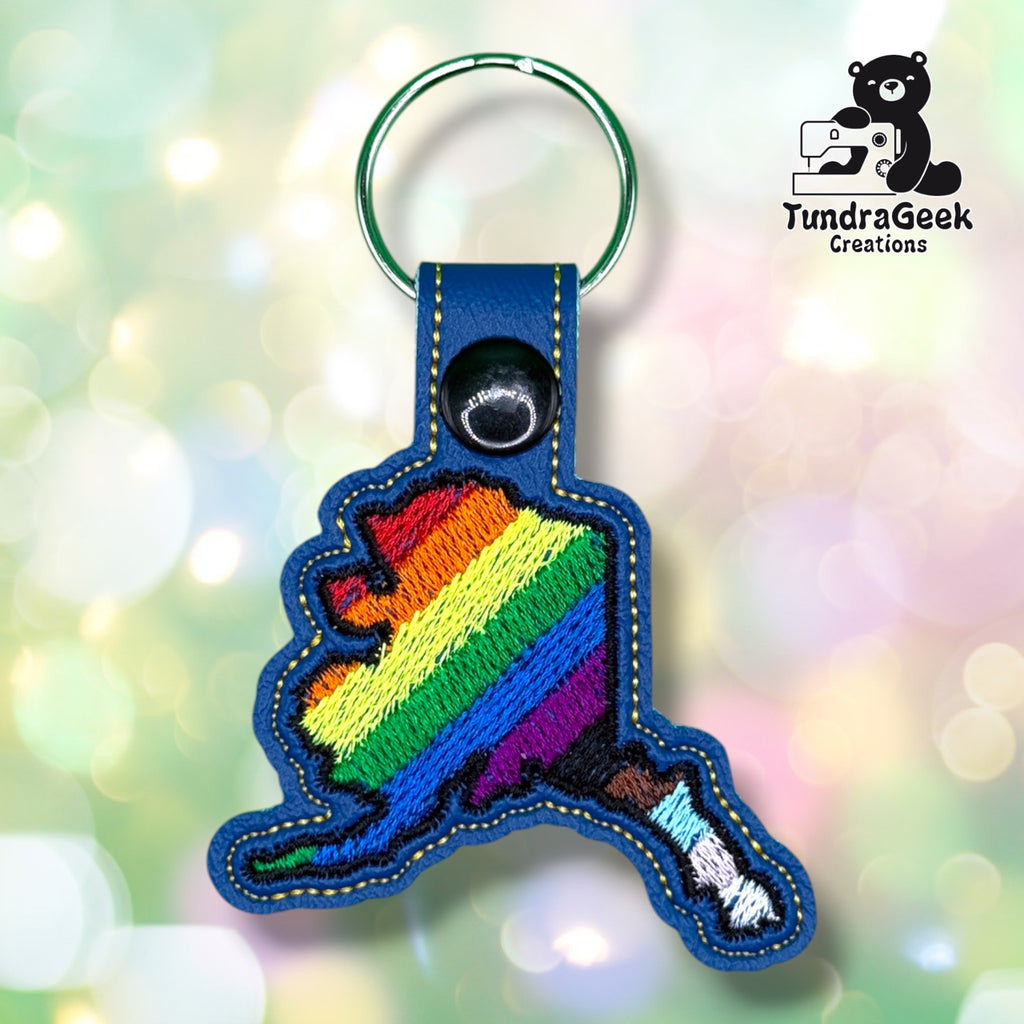 Front view of a blue vinyl keychain shaped like the state of Alaska, embroidered with diagonal rainbow pride stripes across the main body that transition into Progress Pride's brown, light blue, and pink stripes along the Aleutian Islands tail. Finished with a black plastic snap closure and silver split ring.