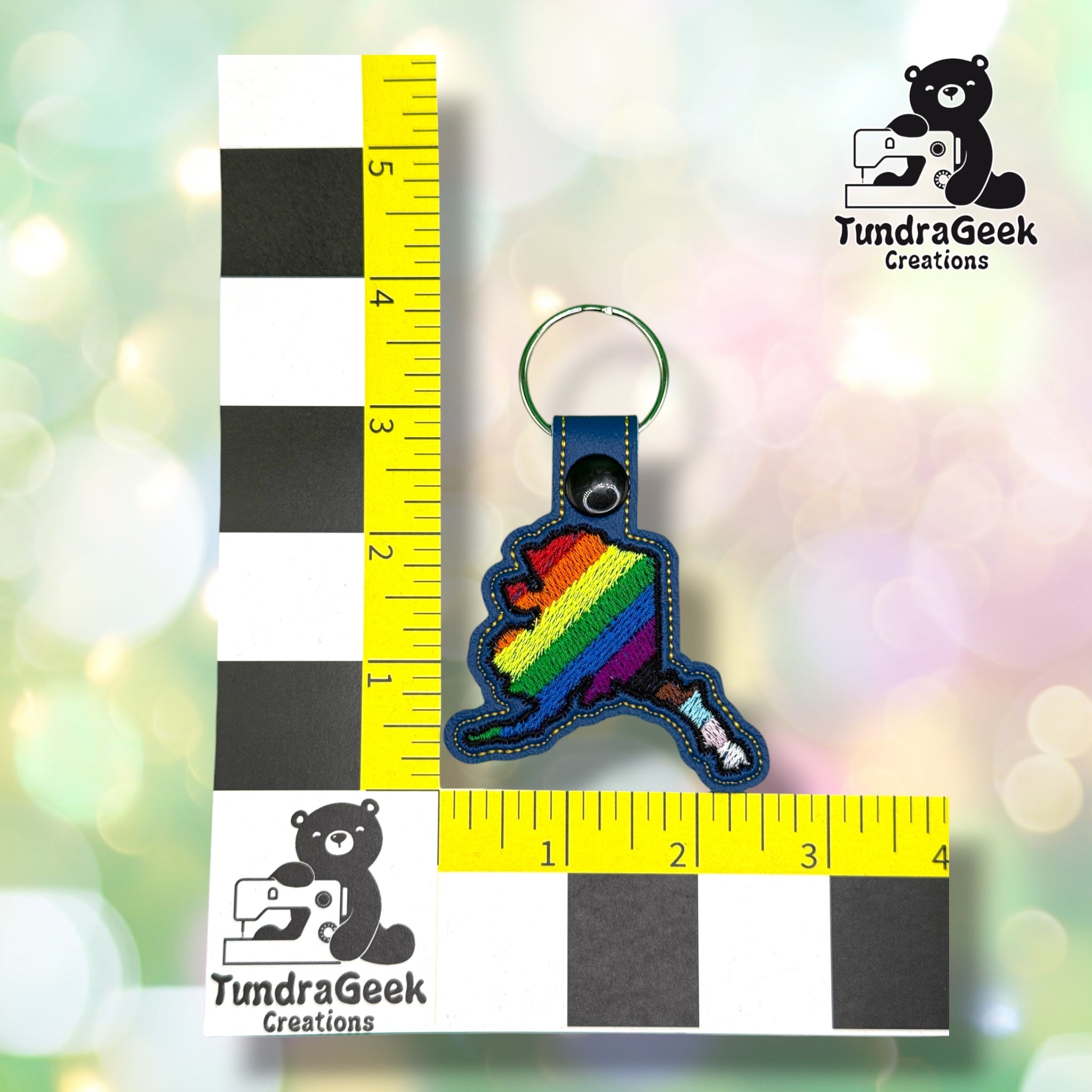 Keychain shown against a black-and-white checkered background with yellow rulers for scale, measuring approximately 3.75 inches tall by 2.5 inches wide including hardware.  ---