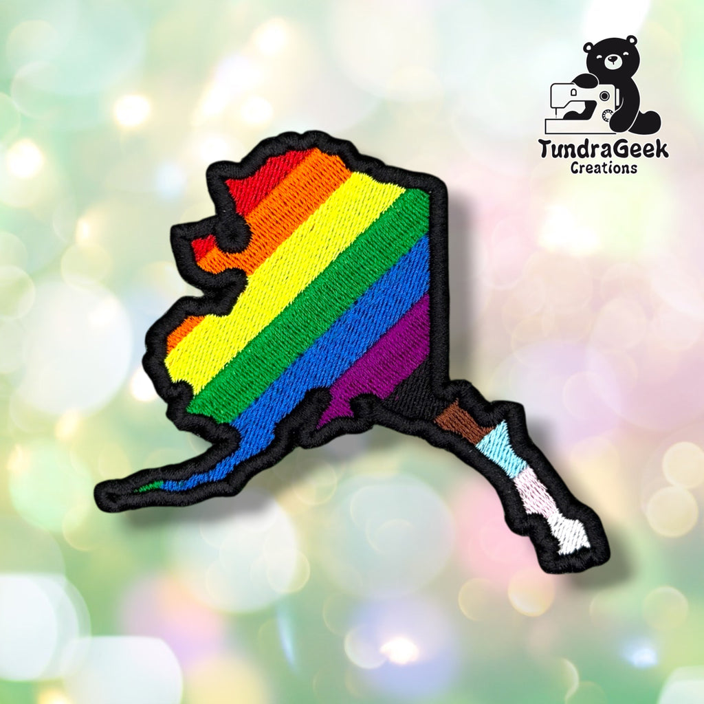 Front view of a patch shaped like the state of Alaska with a black merrowed edge, embroidered with diagonal rainbow stripes across the main body that transition into Progress Pride's brown, light blue, and pink stripes along the Aleutian Islands tail.