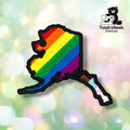 Front view of a patch shaped like the state of Alaska with a black merrowed edge, embroidered with diagonal rainbow stripes across the main body that transition into Progress Pride's brown, light blue, and pink stripes along the Aleutian Islands tail.