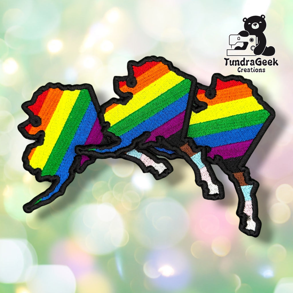 Three Alaska State Pride patches fanned out together, each showing the same rainbow-to-Progress-Pride Alaska state design with black merrowed edges.