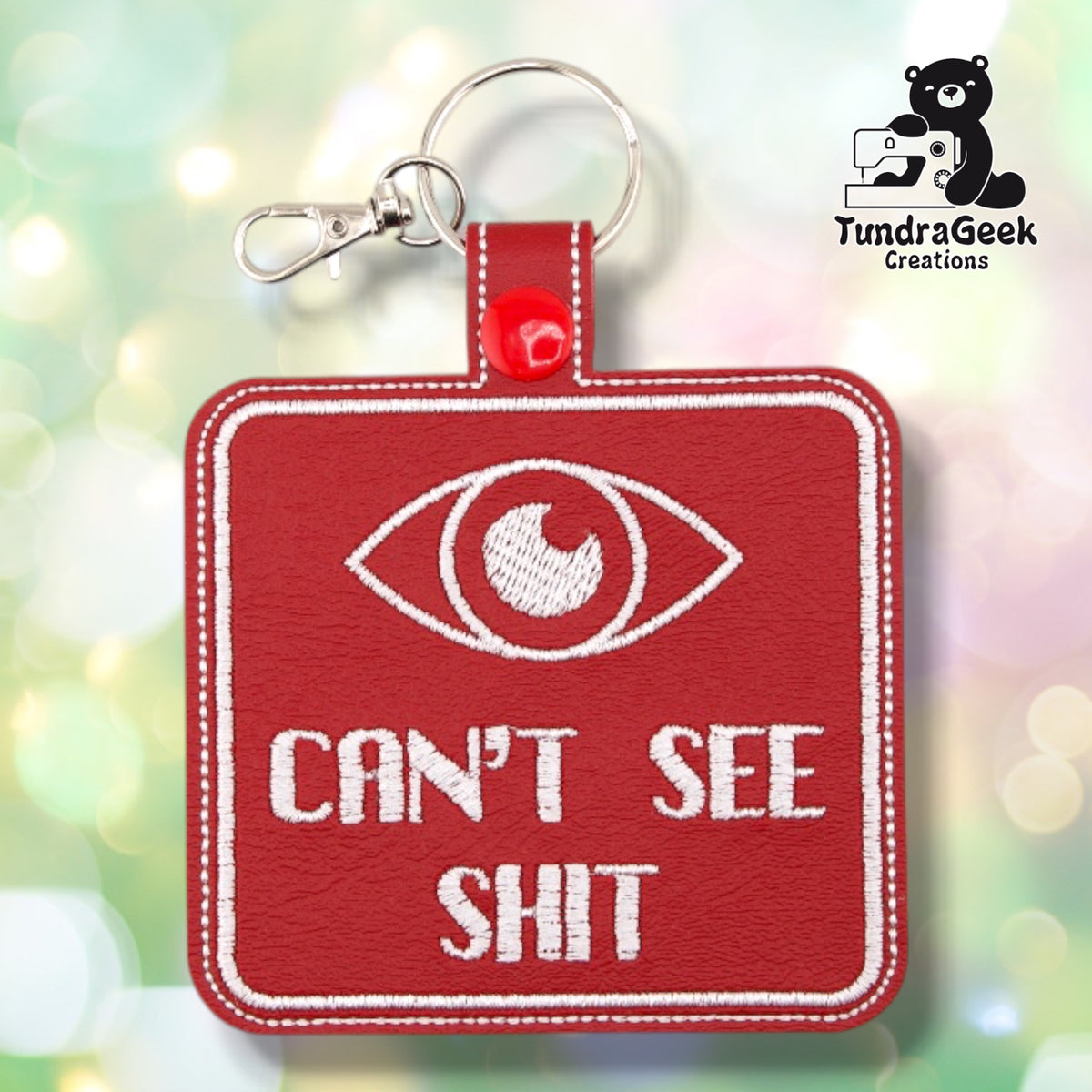 Red square embroidered vinyl keychain badge featuring a white embroidered eye graphic above the text "CAN'T SEE SHIT" in white. White border on a red background. Silver lobster claw clip and red snap hardware at top.