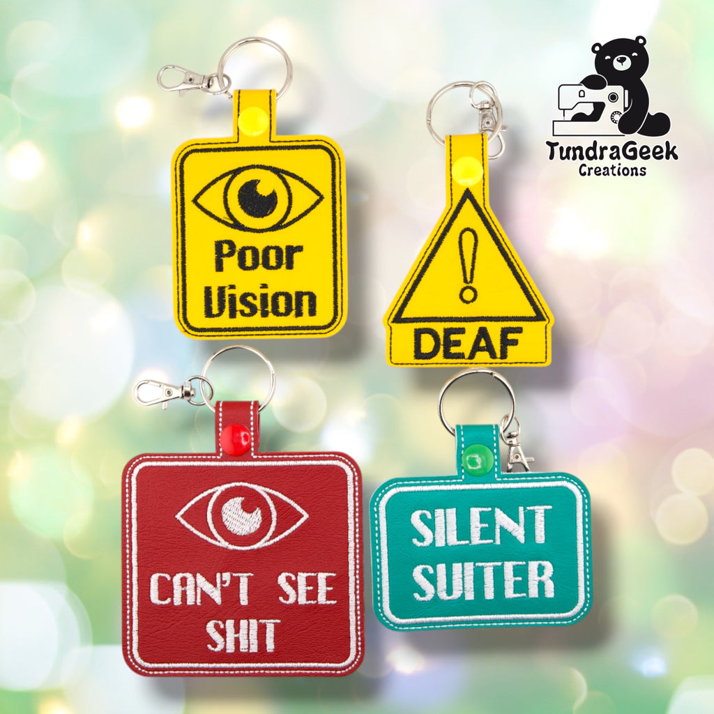 Four embroidered vinyl awareness keychain badges arranged in two rows on a soft bokeh background. Top row: Poor Vision (yellow square), Deaf (yellow triangle). Bottom row: Can't See Shit (red square), Silent Suiter (teal rectangle). All badges shown with silver lobster claw clip hardware. Replaces the prior group shot, which still showed the discontinued green Poor Vision badge.