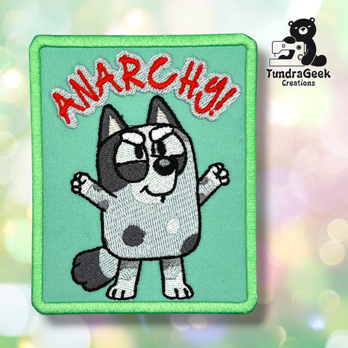Front view of a teal rectangular patch with a green merrowed edge, embroidered with a grey-and-white spotted puppy standing with paws raised, and "ANARCHY!" lettering in red with a white outline above its head.