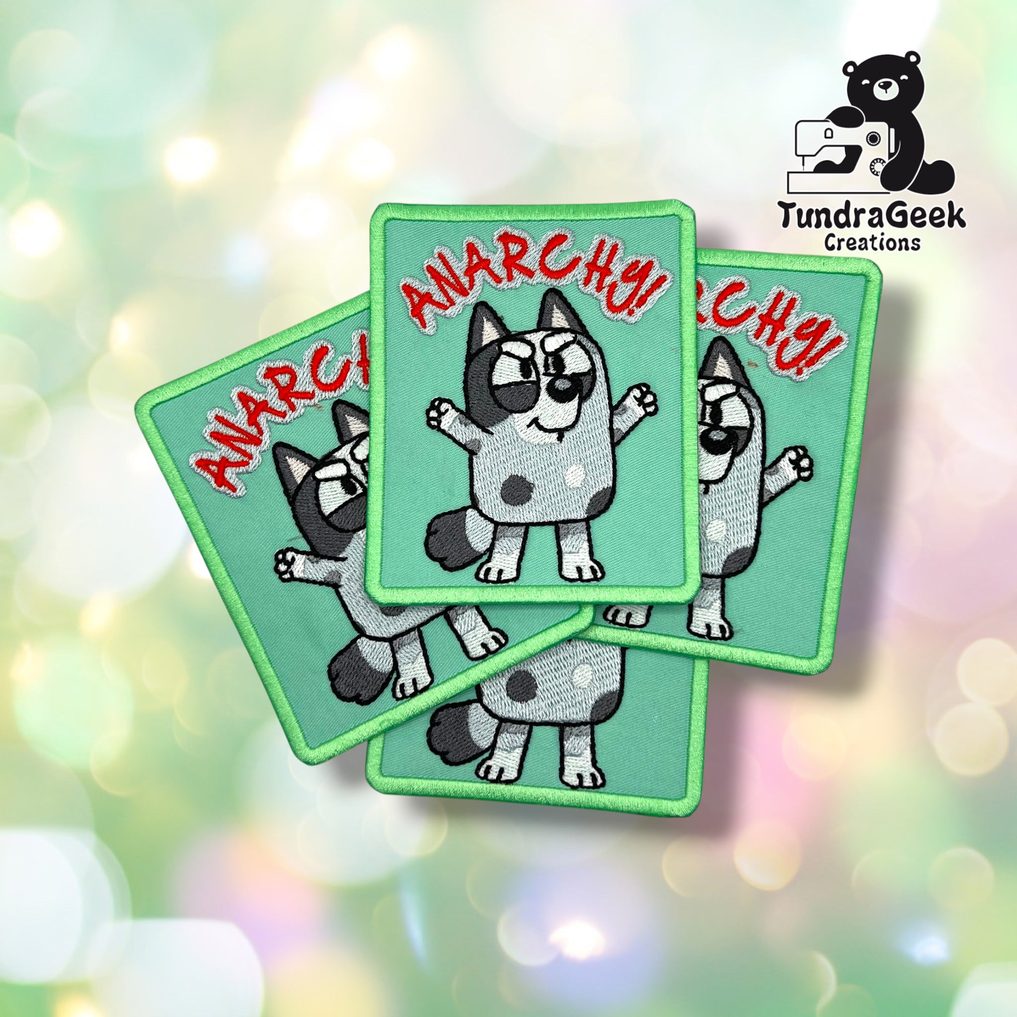 Four green embroidered patches with a cartoon dog, raised arms, sunglasses, and the word “ANARCHY!” in red. “TundraGeek Creations” logo is on the left. Geometric background.