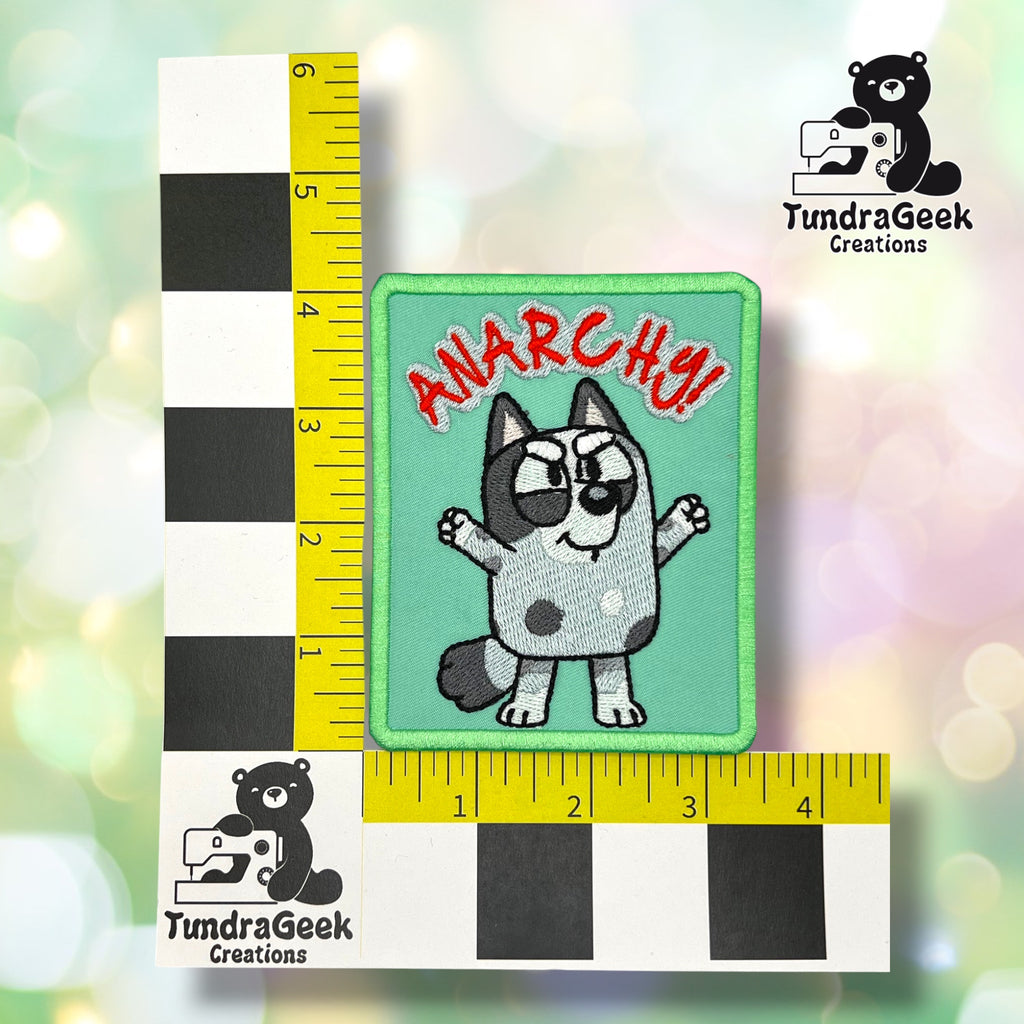 A rectangular embroidered patch featuring a cartoon gray and white cat with the word 'ANARCHY!' in red above it, placed on a blurred pastel background with a black-and-white ruler and yellow tape measure for scale. TundraGeek Creations logos appear in the corners.
