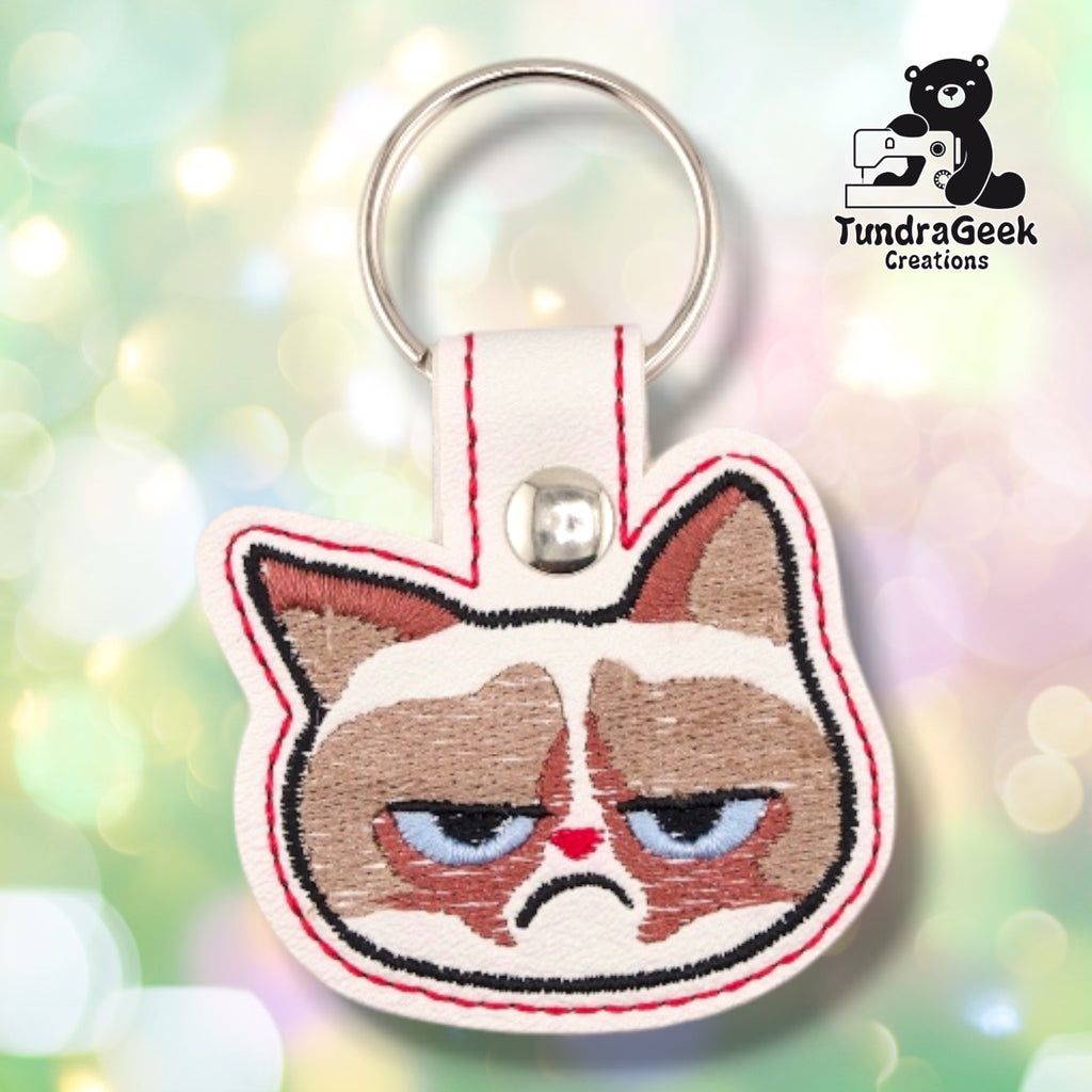 Front of a cream vinyl keychain embroidered with a highly detailed, annoyed-looking cat face — brown and tan markings, heavy-lidded blue eyes, small red nose, and a deep frown. Red merrowed-style edge stitching. Silver snap closure and metal split ring at top.