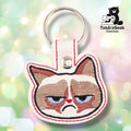 Front of a cream vinyl keychain embroidered with a highly detailed, annoyed-looking cat face — brown and tan markings, heavy-lidded blue eyes, small red nose, and a deep frown. Red merrowed-style edge stitching. Silver snap closure and metal split ring at top.