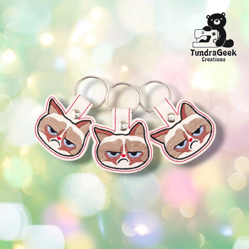 Three identical Annoyed Kitty keychains fanned out together, each showing the same detailed annoyed cat face and red edge stitching.