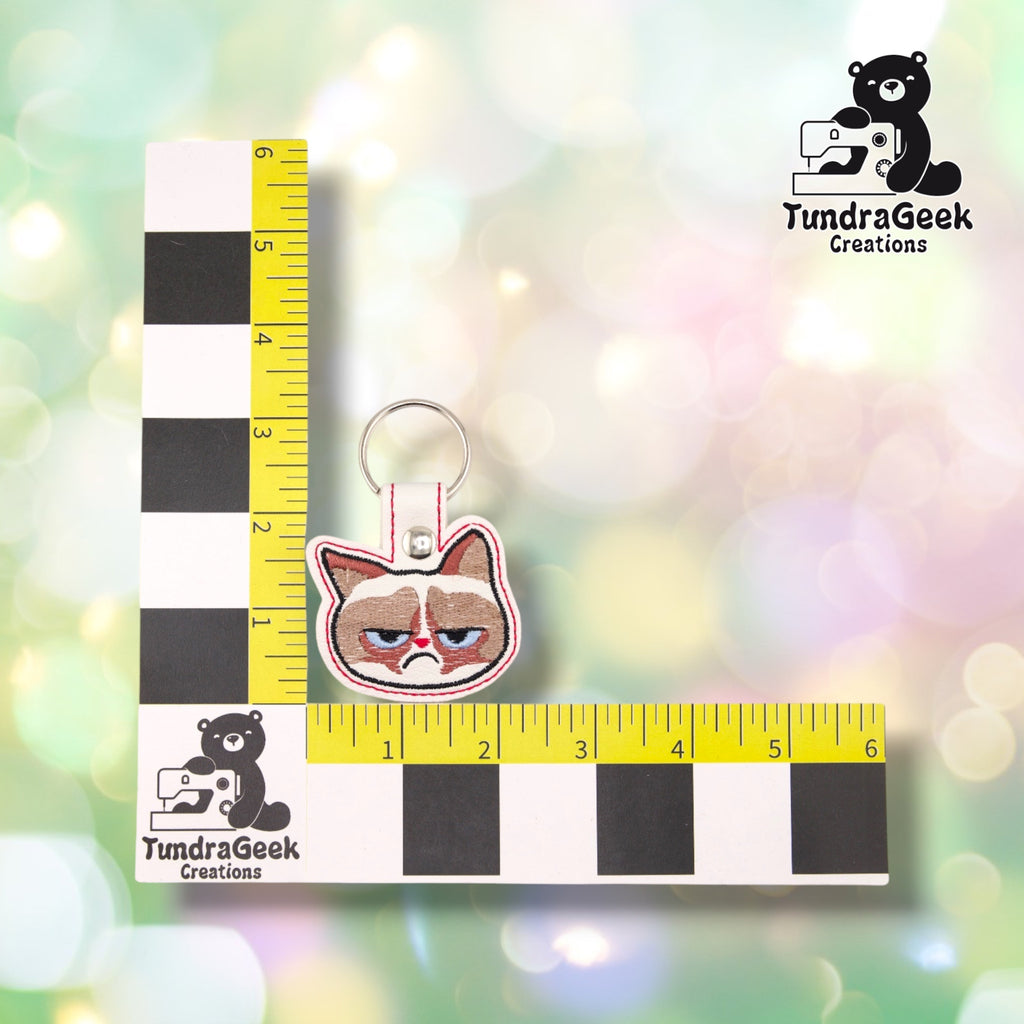 Annoyed Kitty keychain shown against a black-and-white checkered background with a yellow ruler for scale, measuring approximately 3.25 inches tall by 2.25 inches wide.  ---