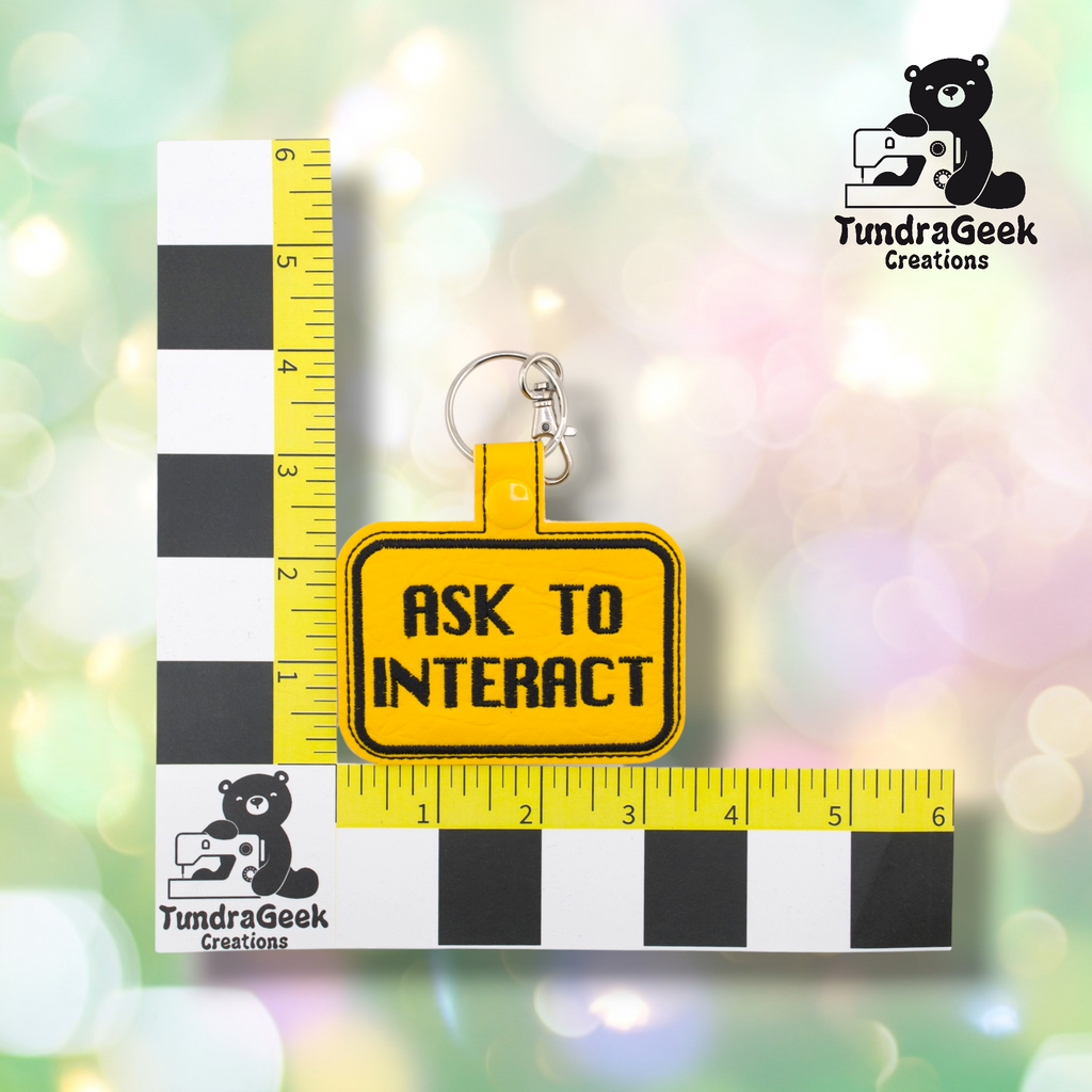 Yellow embroidered vinyl keychain badge reading “ASK TO INTERACT” in black text with a black border. Shown against a ruler indicating approximately 3 inches wide and 2 inches tall. Silver lobster claw clip and yellow snap hardware at top.