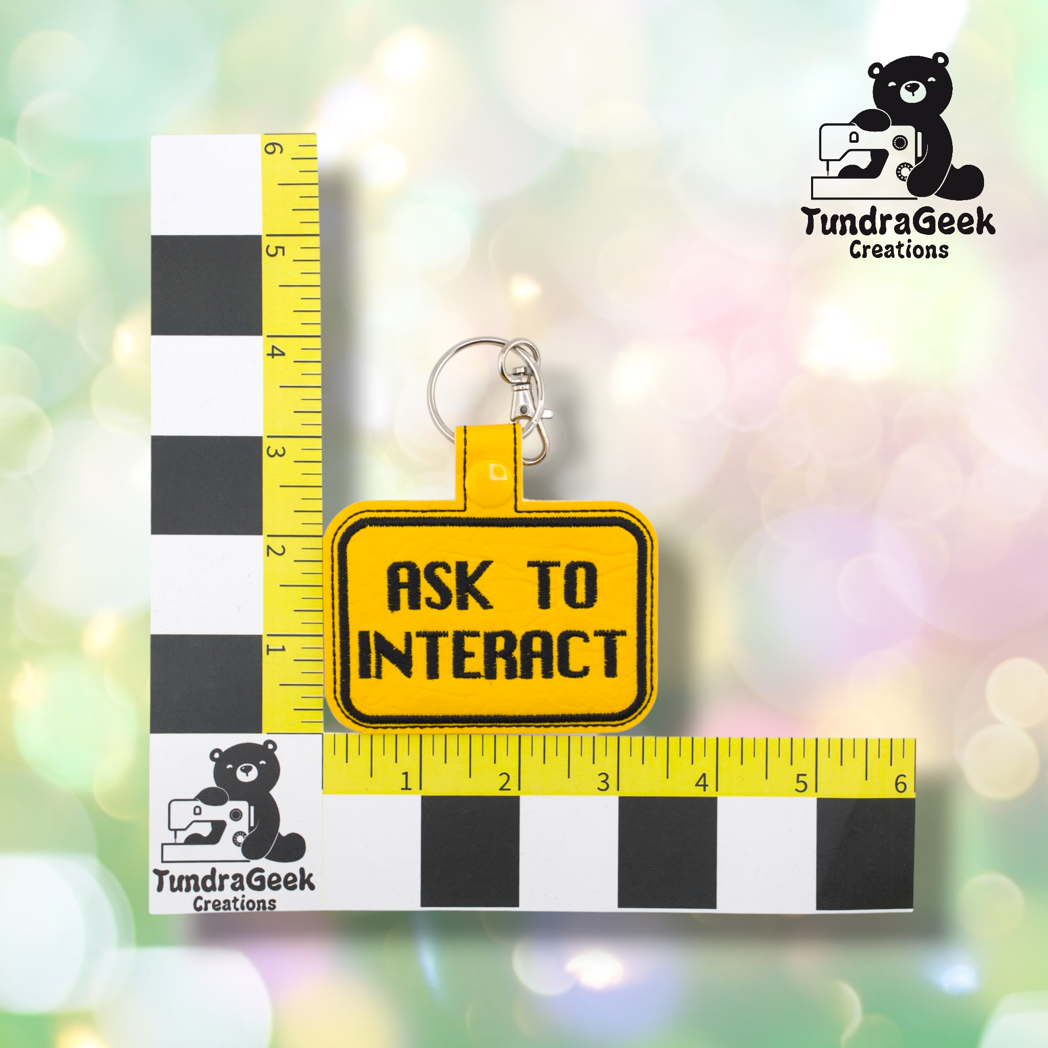 Yellow embroidered vinyl keychain badge reading “ASK TO INTERACT” in black text with a black border. Shown against a ruler indicating approximately 3 inches wide and 2 inches tall. Silver lobster claw clip and yellow snap hardware at top.