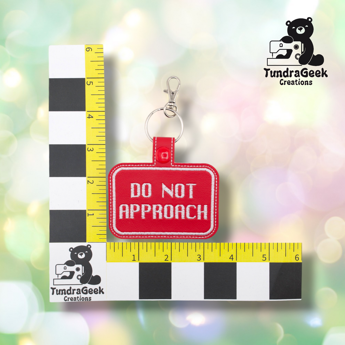 A red rectangular vinyl fabric keychain with white embroidered text reading 'DO NOT APPROACH' and a silver clasp, shown next to a black-and-white measuring block and yellow ruler for scale on a soft pastel bokeh background. TundraGeek Creations logo appears in the top right and bottom left.