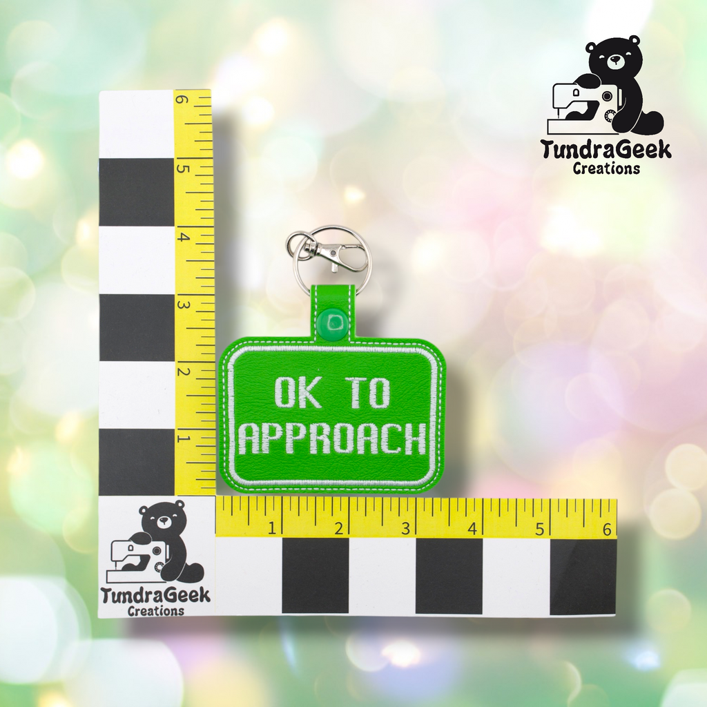 Bright green embroidered vinyl keychain badge reading “OK TO APPROACH” in white text with a white border. Shown against a ruler indicating approximately 3 inches wide and 2 inches tall. Silver lobster claw clip and green snap hardware at top.