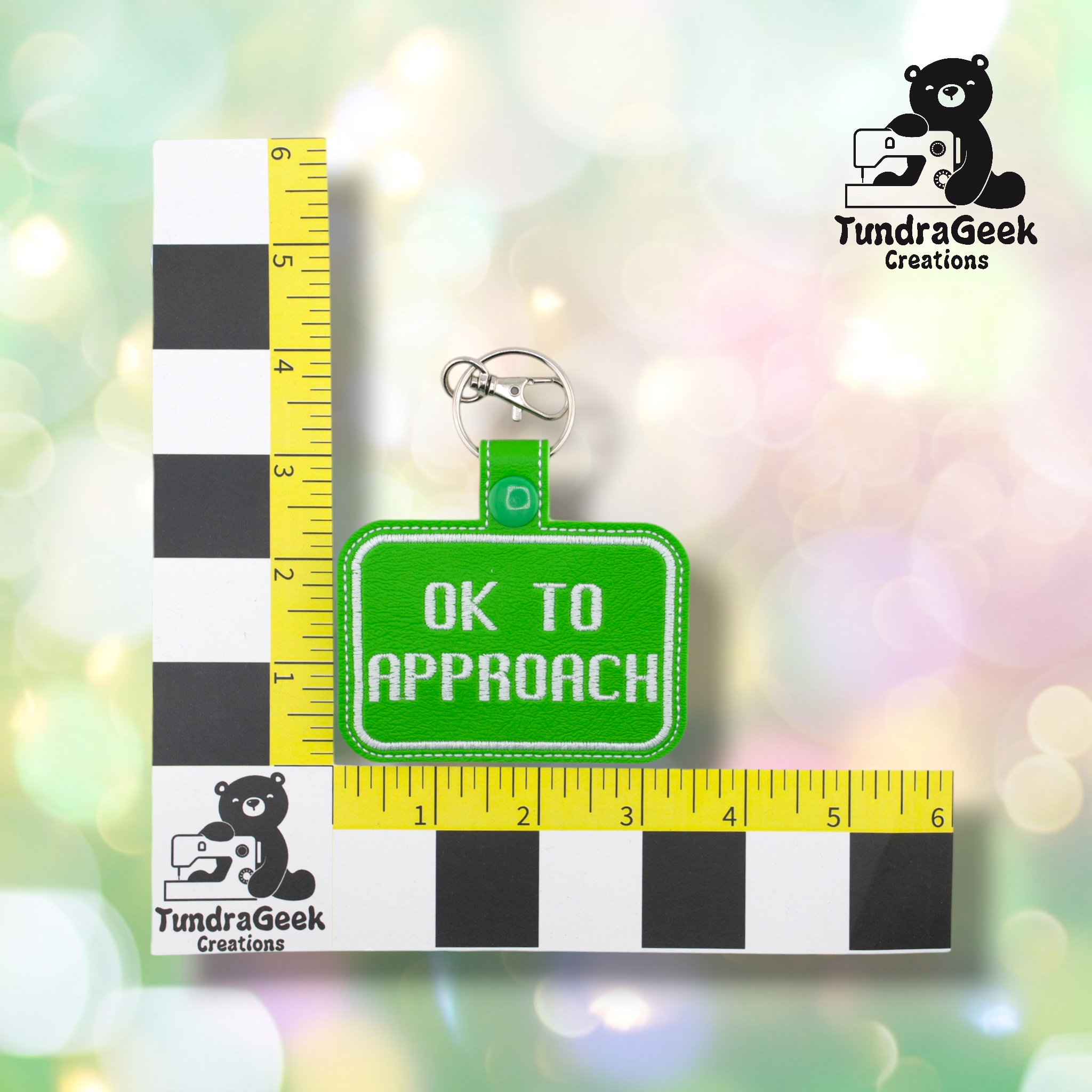 Bright green embroidered vinyl keychain badge reading “OK TO APPROACH” in white text with a white border. Shown against a ruler indicating approximately 3 inches wide and 2 inches tall. Silver lobster claw clip and green snap hardware at top.