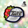 Front view of a baseball-cap-shaped patch embroidered with "DON'T BE AN ASSHAT" in black block lettering across a light blue cap with navy trim, accented by a red and pink tattoo-style flower with green leaves on the left side.
