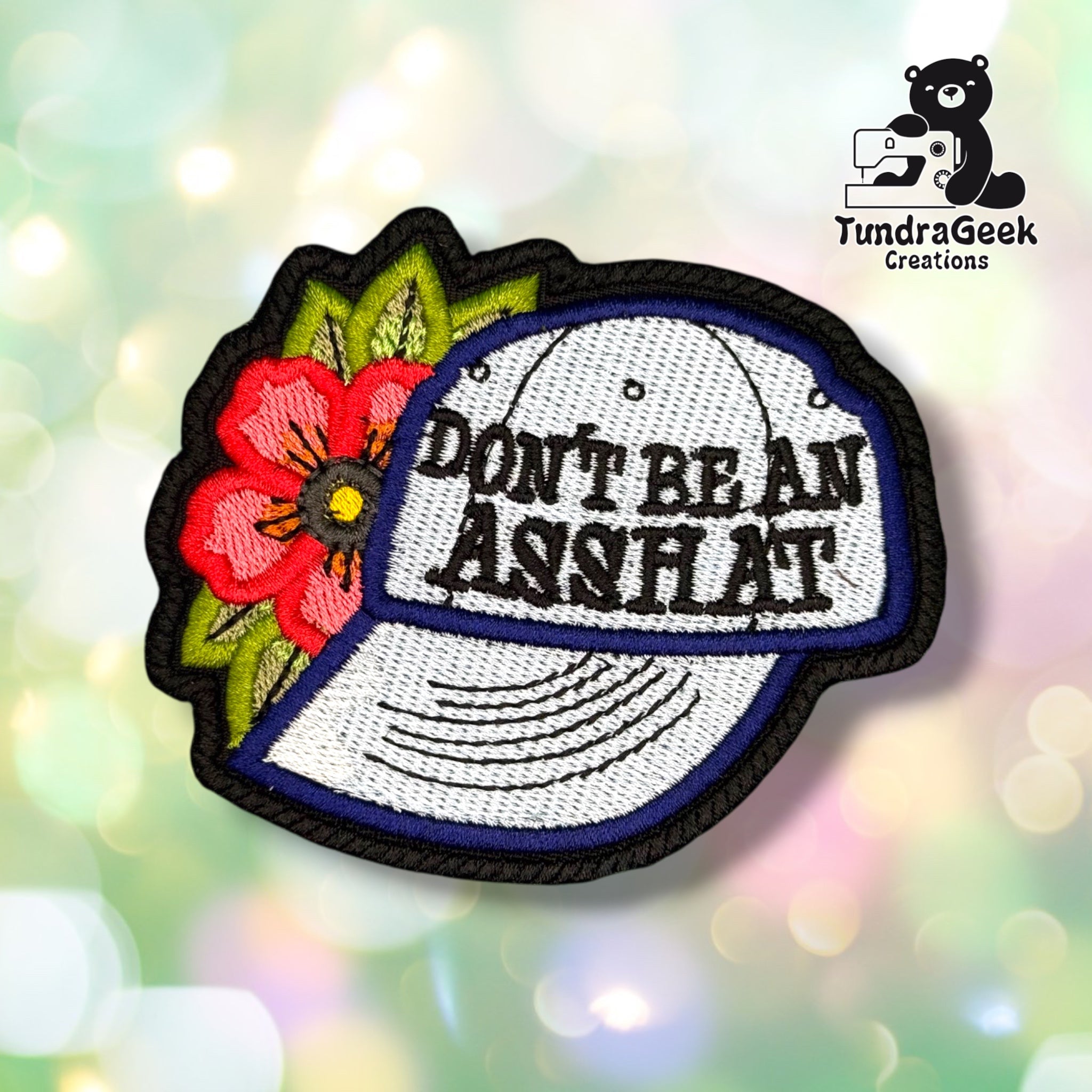 Front view of a baseball-cap-shaped patch embroidered with "DON'T BE AN ASSHAT" in black block lettering across a light blue cap with navy trim, accented by a red and pink tattoo-style flower with green leaves on the left side.