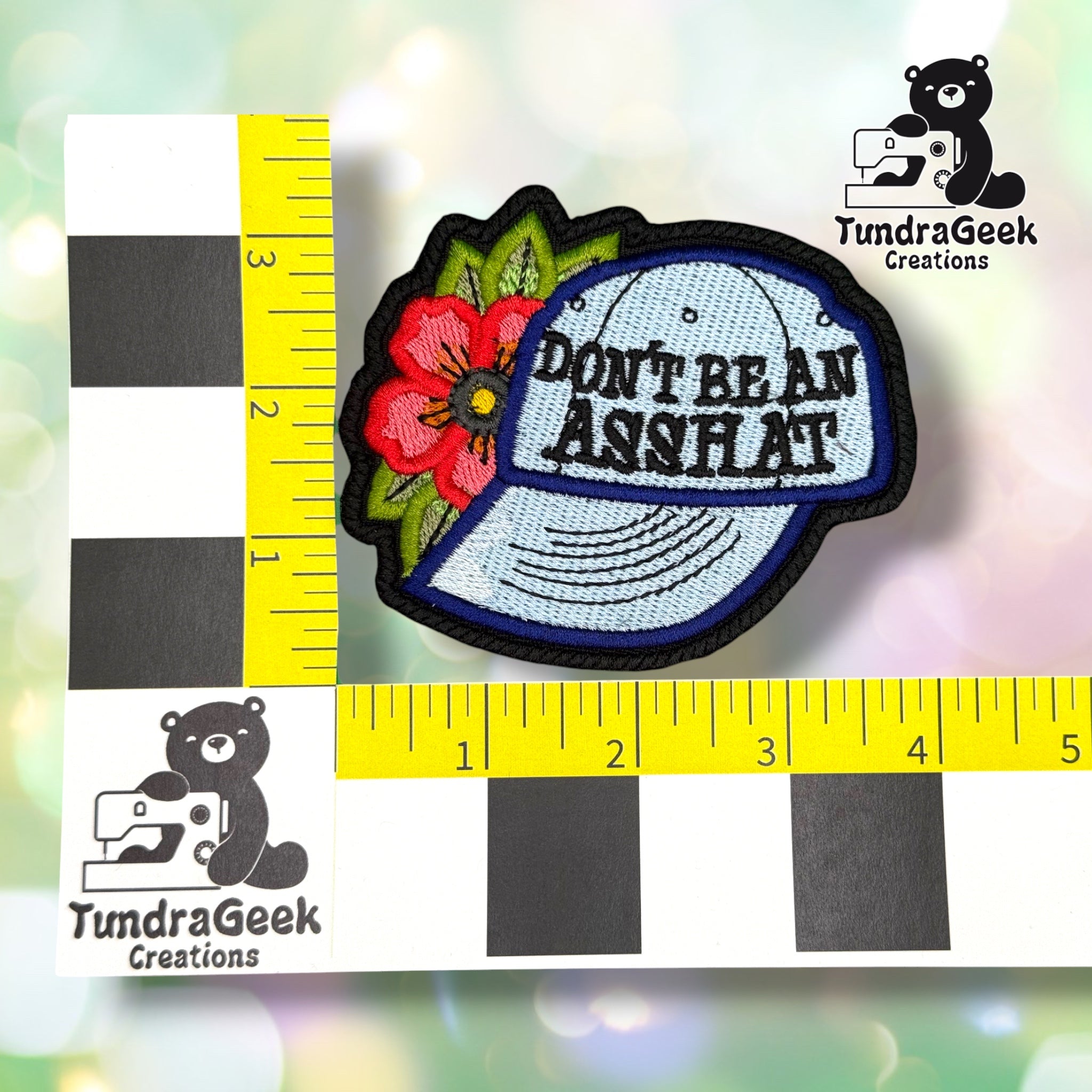 Patch shown against a black-and-white checkered background with yellow rulers for scale, measuring approximately 4 inches wide by 3.5 inches tall.  ---