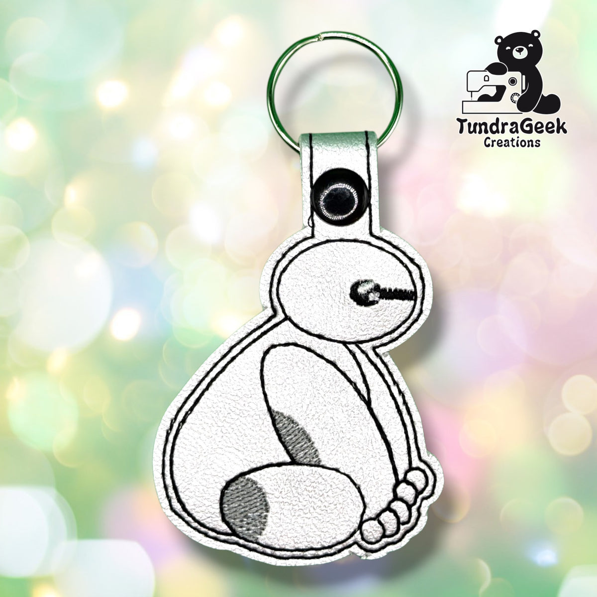Front view of a white vinyl keychain shaped like a rounded robot companion sitting upright, with grey oval shading on its body, holding a black ball-tipped wand in one hand. Finished with a black plastic snap closure and green split ring.