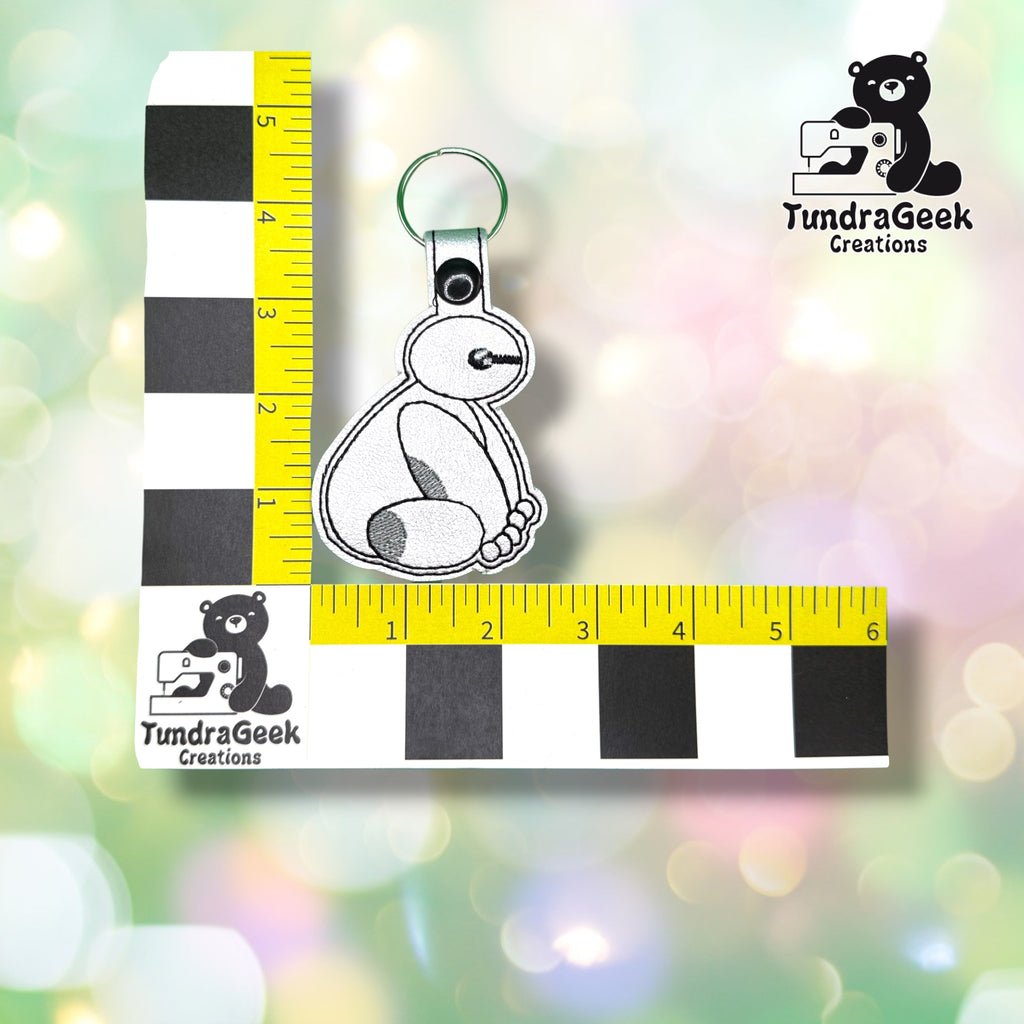Keychain shown against a black-and-white checkered background with yellow rulers for scale, measuring approximately 2.5 inches wide by 4.75 inches tall, including hardware.  ---