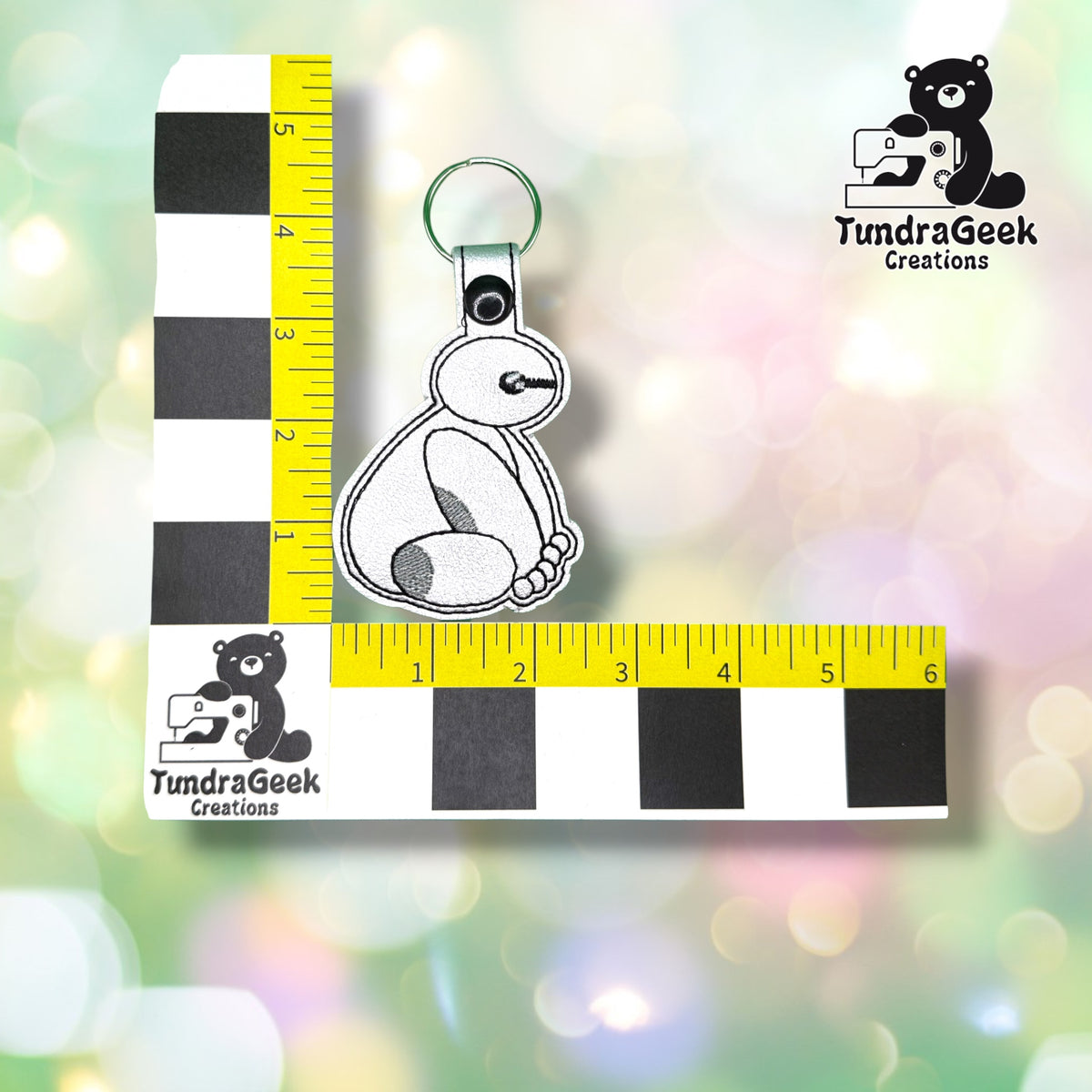 Keychain shown against a black-and-white checkered background with yellow rulers for scale, measuring approximately 2.5 inches wide by 4.75 inches tall, including hardware.  ---