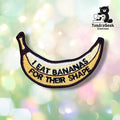 Front view of a banana-shaped patch embroidered in yellow, white, and brown, with "I EAT BANANAS FOR THEIR SHAPE" stitched in black block lettering along the curve of the banana.
