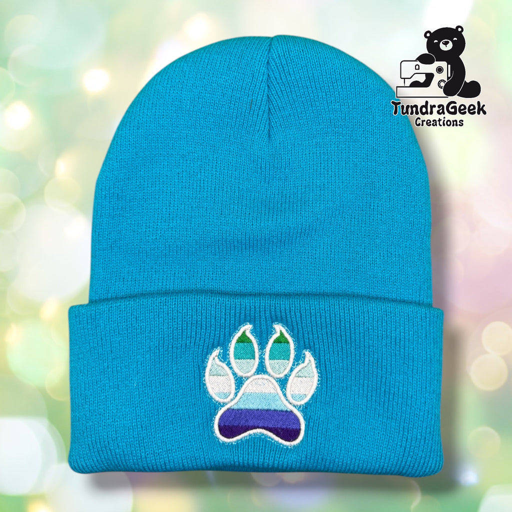 Neon blue cuffed acrylic beanie with a white-outlined paw print embroidered on the cuff. The toe beans and main pad display MLM pride flag stripes in shades of green, teal, light blue, and purple. TundraGeek Creations logo in upper right corner.