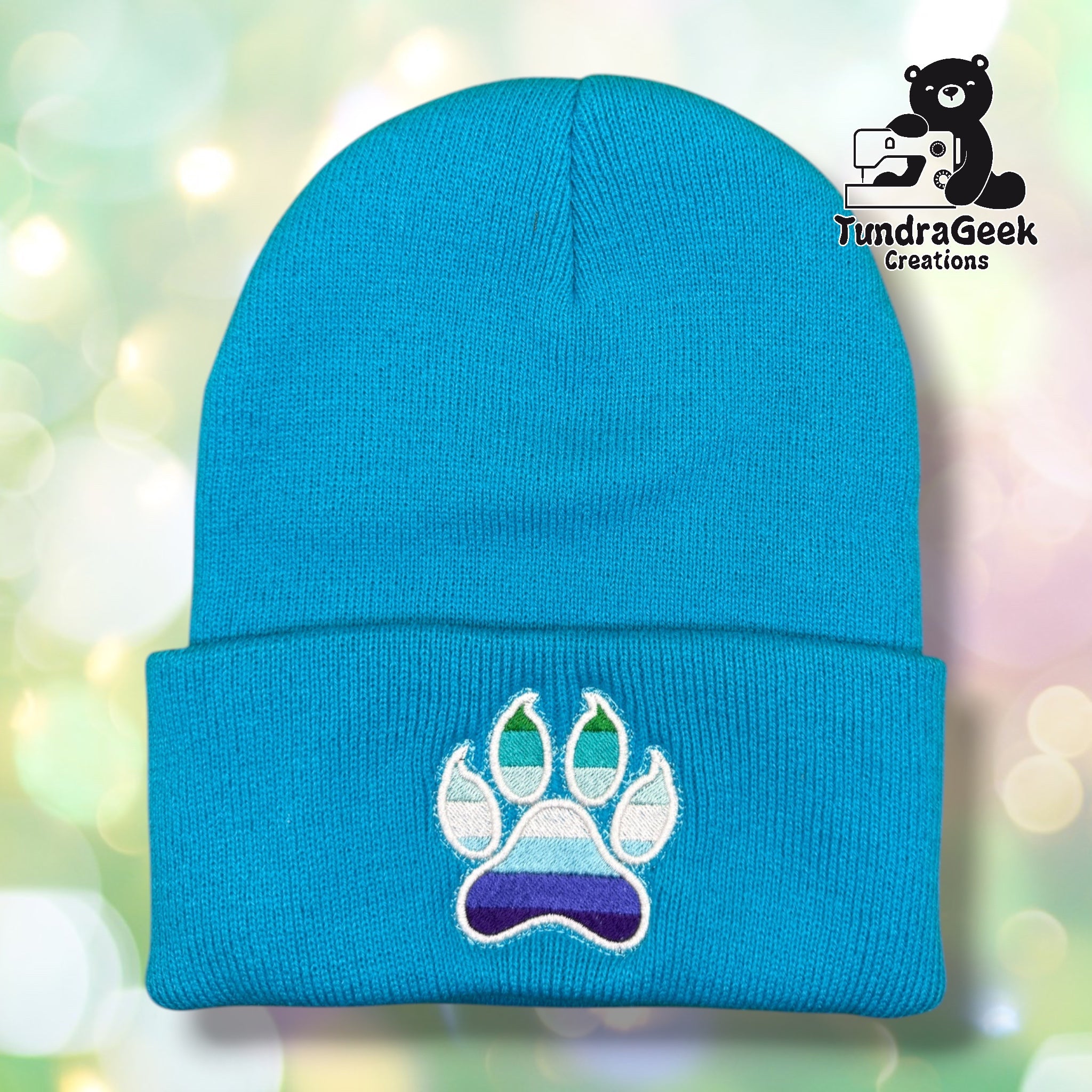 Neon blue cuffed acrylic beanie with a white-outlined paw print embroidered on the cuff. The toe beans and main pad display MLM pride flag stripes in shades of green, teal, light blue, and purple. TundraGeek Creations logo in upper right corner.