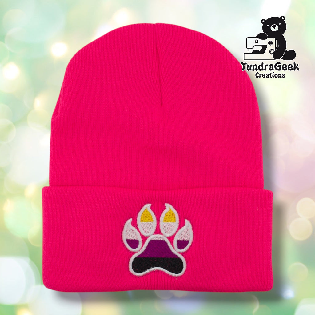 Neon pink cuffed acrylic beanie with a white-outlined paw print embroidered on the cuff. The toe beans and main pad are filled with nonbinary pride flag colors — yellow, white, purple, and black stripes. TundraGeek Creations logo in upper right corner.