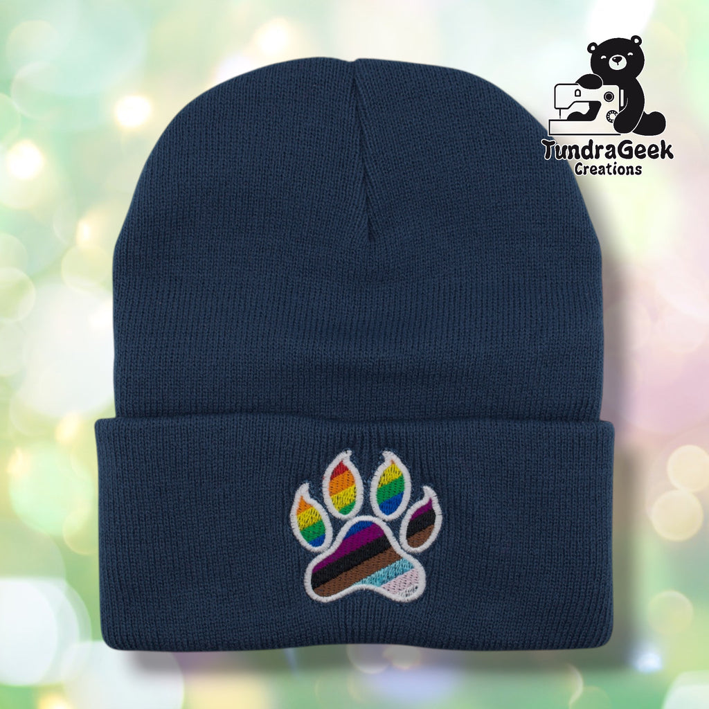 Millennium Blue cuffed acrylic beanie with a white-outlined paw print embroidered on the cuff. The toe beans display rainbow stripes; the main pad features Progress Pride flag stripes including black, brown, light blue, pink, and white. TundraGeek Creations logo in upper right corner.