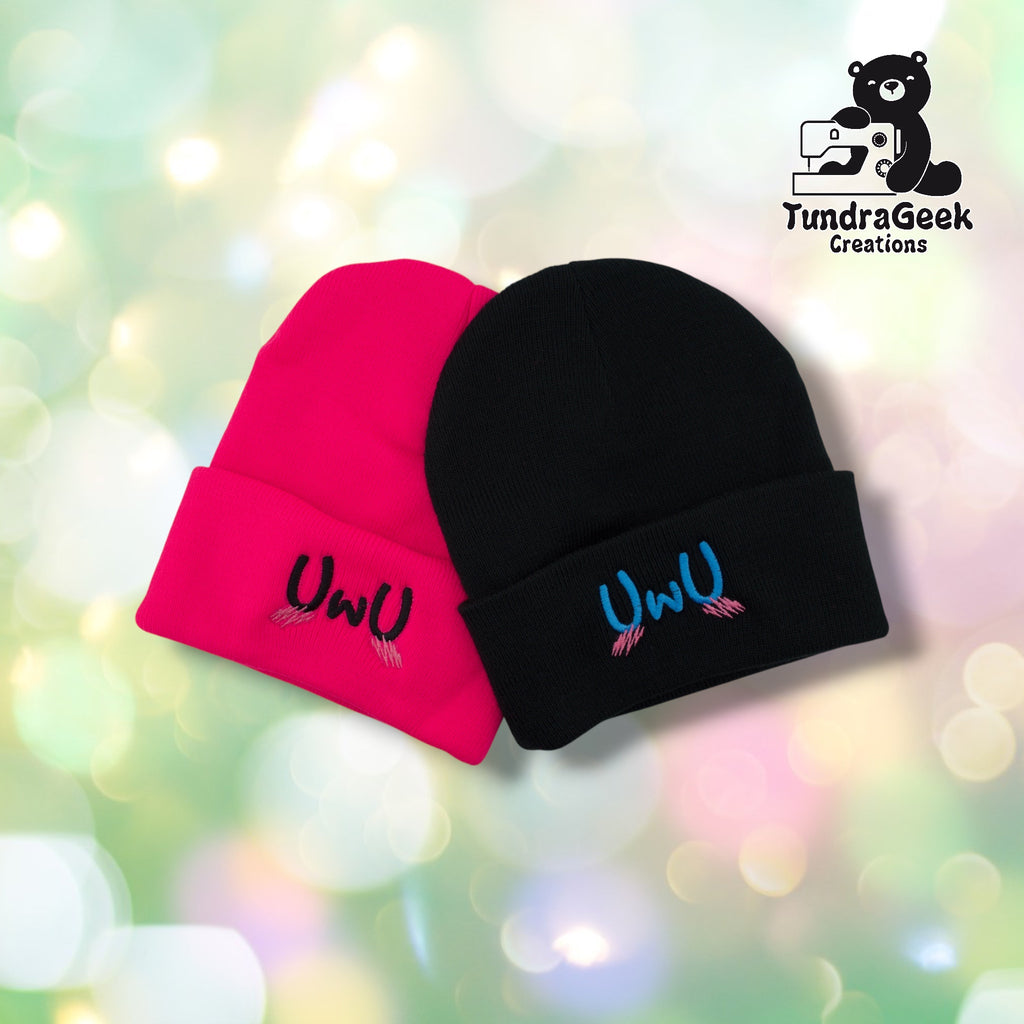 lack and neon pink UwU beanies side by side on a pastel bokeh background. Both feature the UwU emoticon embroidered on the cuff with pink blush marks beneath. TundraGeek Creations logo in upper right corner.
