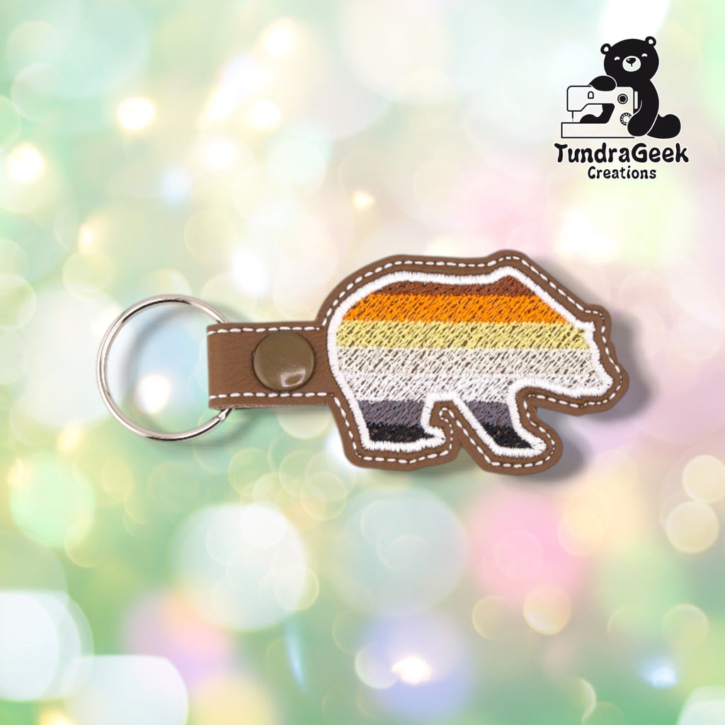 Front of a bear-shaped vinyl keychain embroidered with the Bear Pride flag stripes (brown, orange, yellow, white, gray, black) from top to bottom. Brown vinyl edge with white outline stitching. Olive snap closure and metal split ring at top.