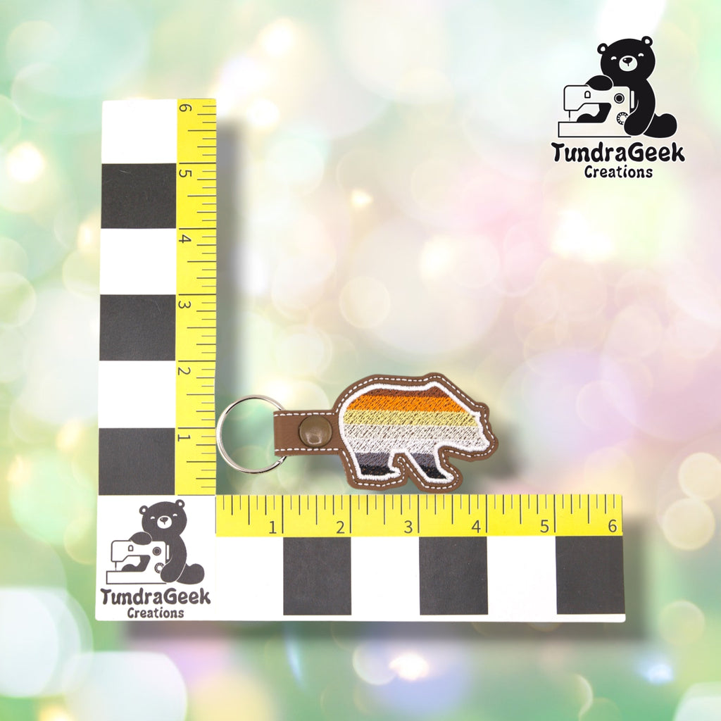 Bear-Shaped Bear Pride keychain shown against a black-and-white checkered background with a yellow ruler for scale, measuring approximately 4.25 inches wide by 1.75 inches tall.  ---