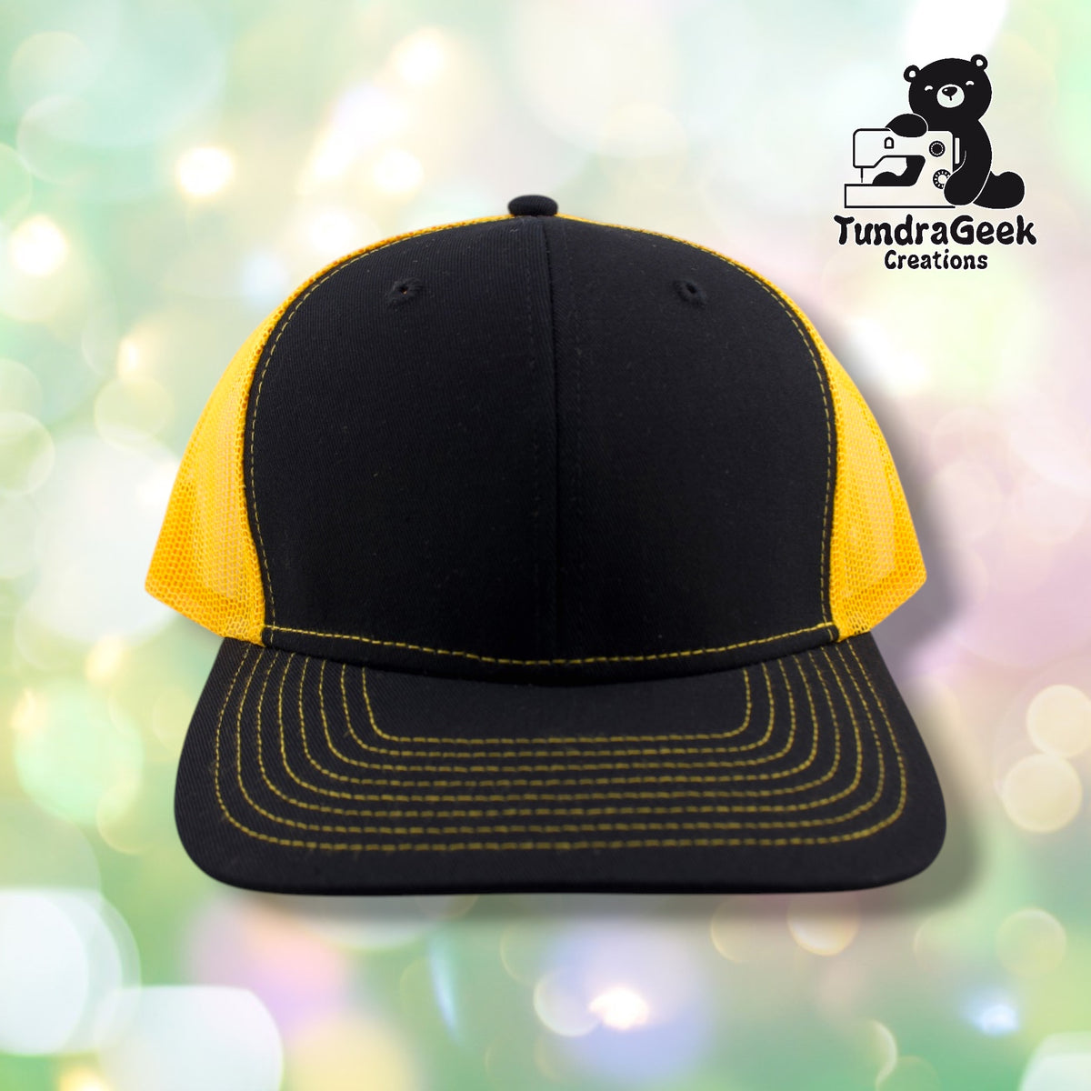 A black and yellow trucker cap with mesh side panels, gold stitching on the bill, displayed against a soft bokeh background with the TundraGeek Creations logo.