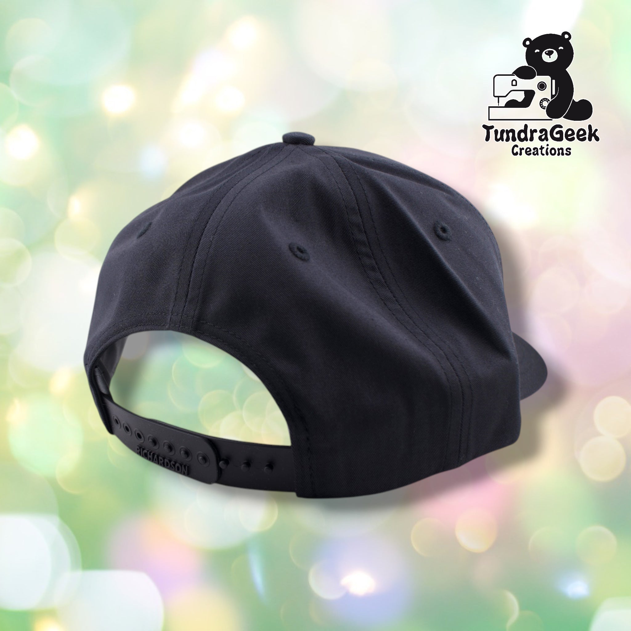 A black adjustable snapback baseball cap displayed against a soft, blurred pastel background with the TundraGeek Creations logo visible in the upper right corner.