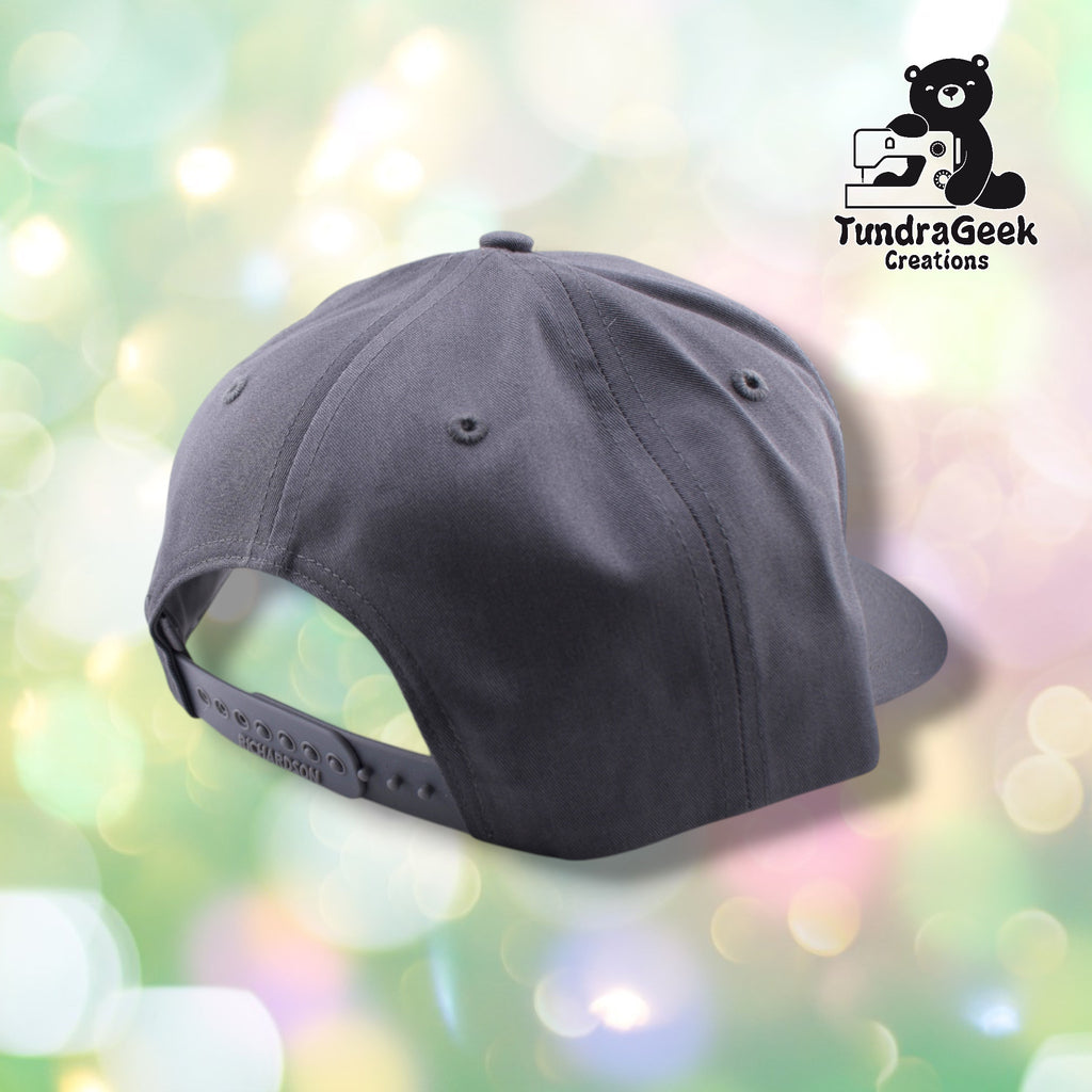A dark gray snapback baseball cap with adjustable back strap displayed against a soft, blurred pastel background with bokeh lighting.